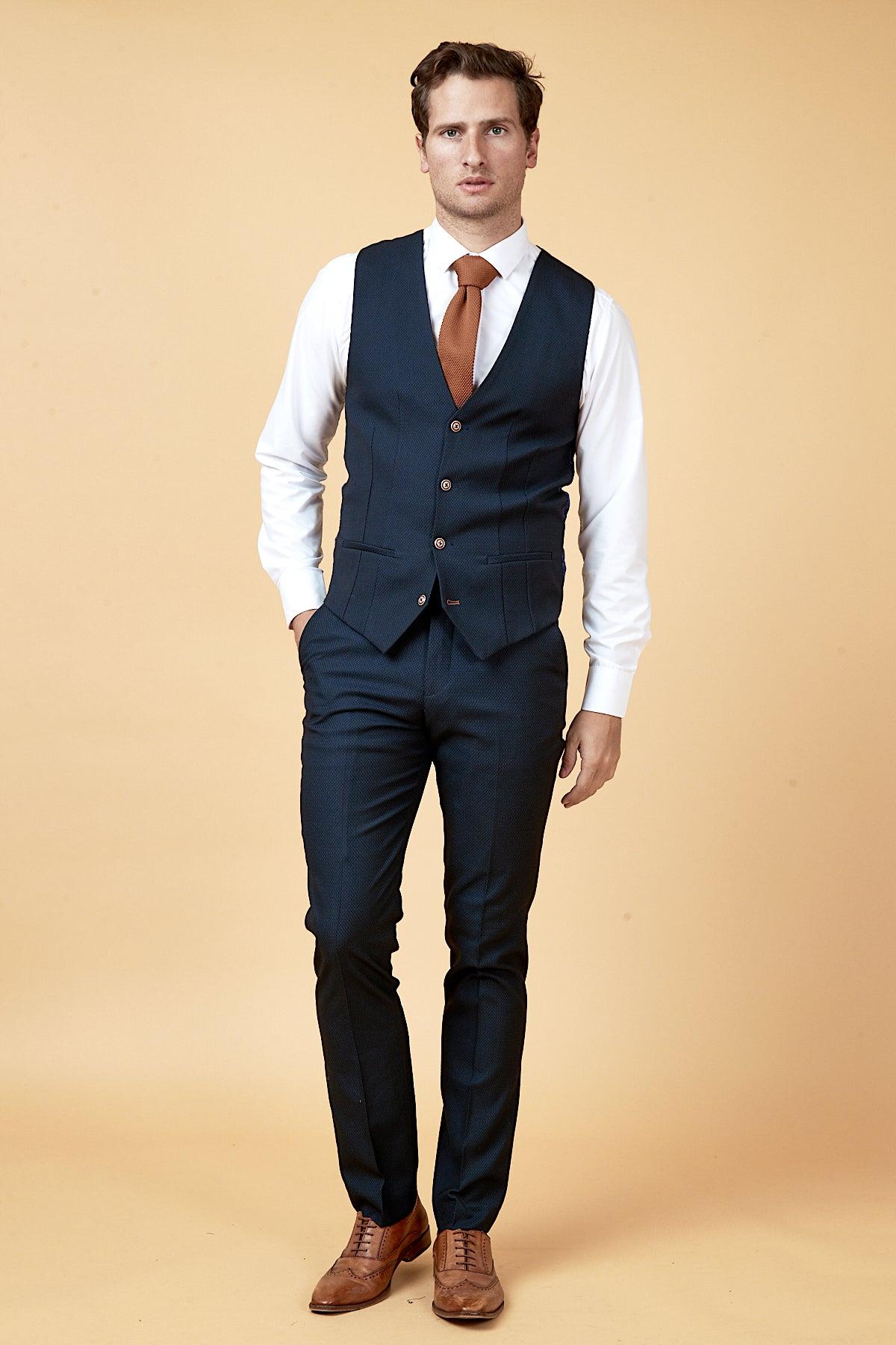A man stands against a beige background wearing the marcdarcy WHU Collection - MAX Navy Three Piece Suit as worn by Max Kilman, paired with a white shirt, brown tie, and dress shoes. One hand is in his pocket as he looks forward neutrally.