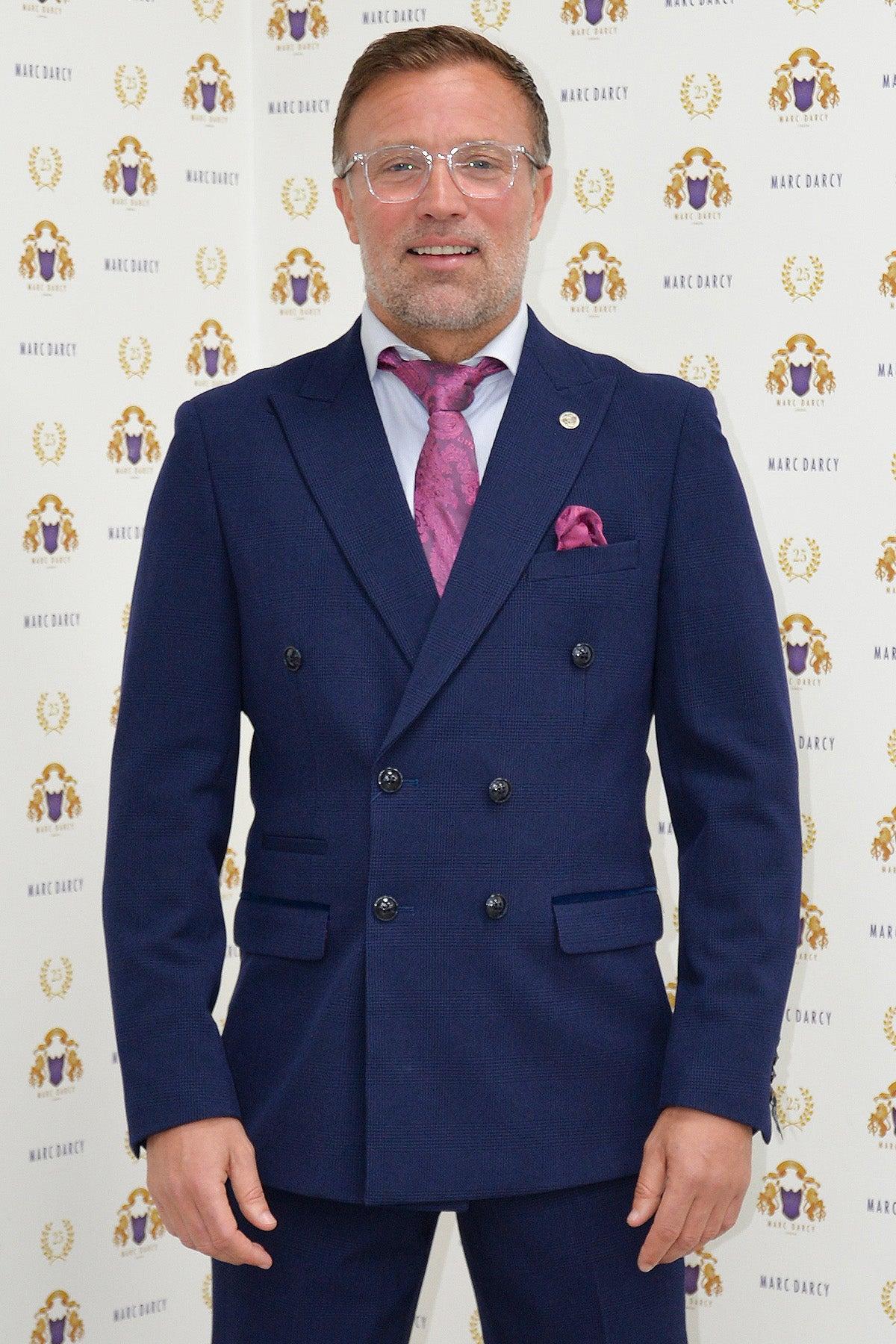 Former Sunderland footballer Michael Gray smiles at the camera in front of a Marc Darcy backdrop, wearing the Bromley Navy Double-Breasted Suit by Marc Darcy, paired with a pink tie and glasses.
