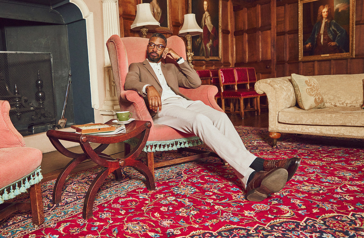 A man in a suit sits relaxed in a pink armchair in a wood-paneled room with portraits, a fireplace, and antique furniture. A green teacup, saucer, and books rest on a small table beside him.