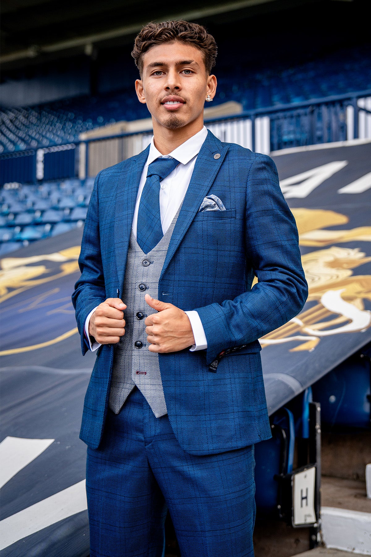 Jeremy Sarmiento stands confidently in a stadium, wearing the marcdarcy WBA Collection - JERRY Blue Suit with Jerry Grey Waistcoat, set against empty blue seats and a large stadium graphic in the background.