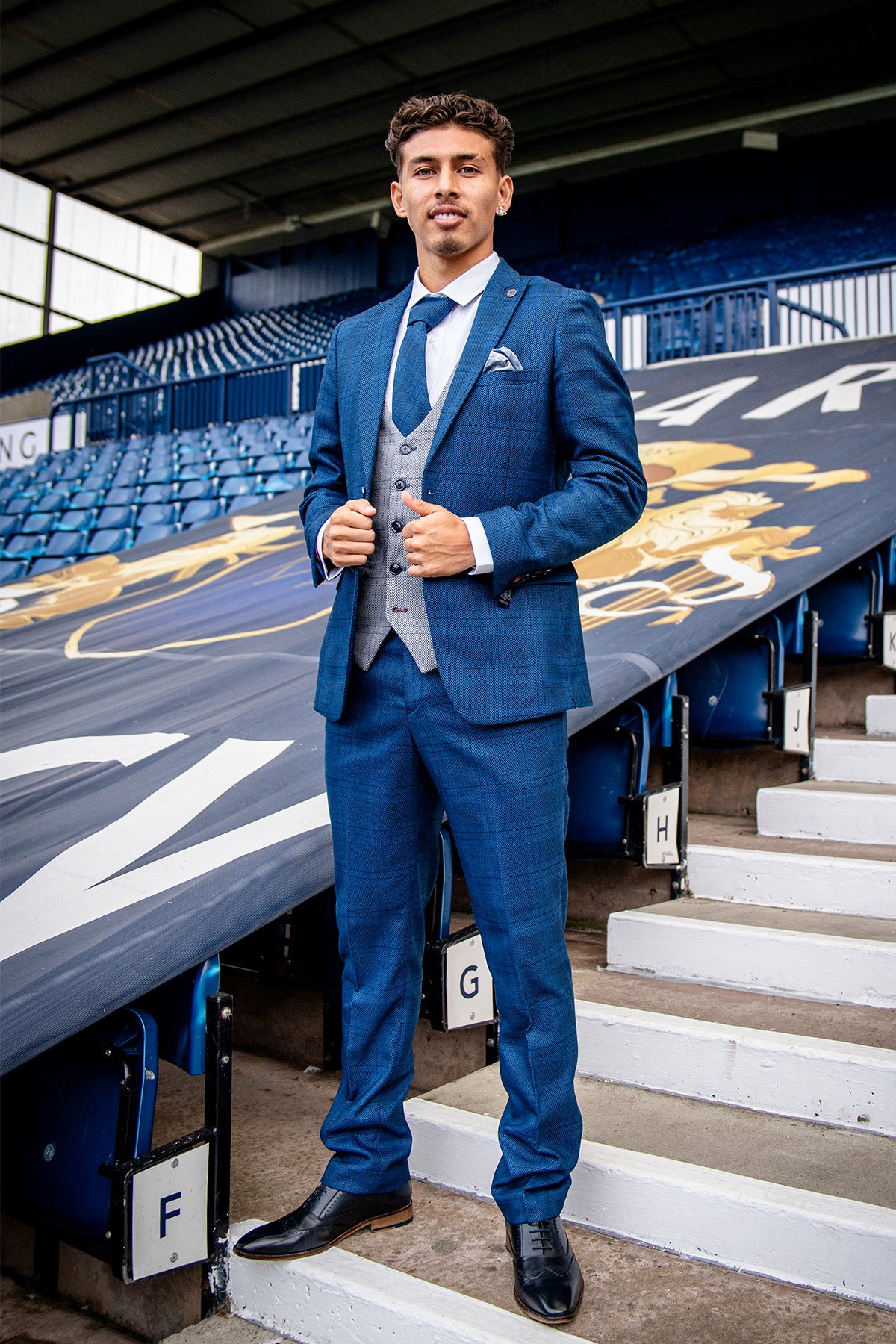 A young man in marcdarcy’s WBA Collection - JERRY Blue Suit with Jerry Grey Waistcoat, as worn by Jeremy Sarmiento, stands in a stadium smiling and adjusting his jacket with a double back vent.