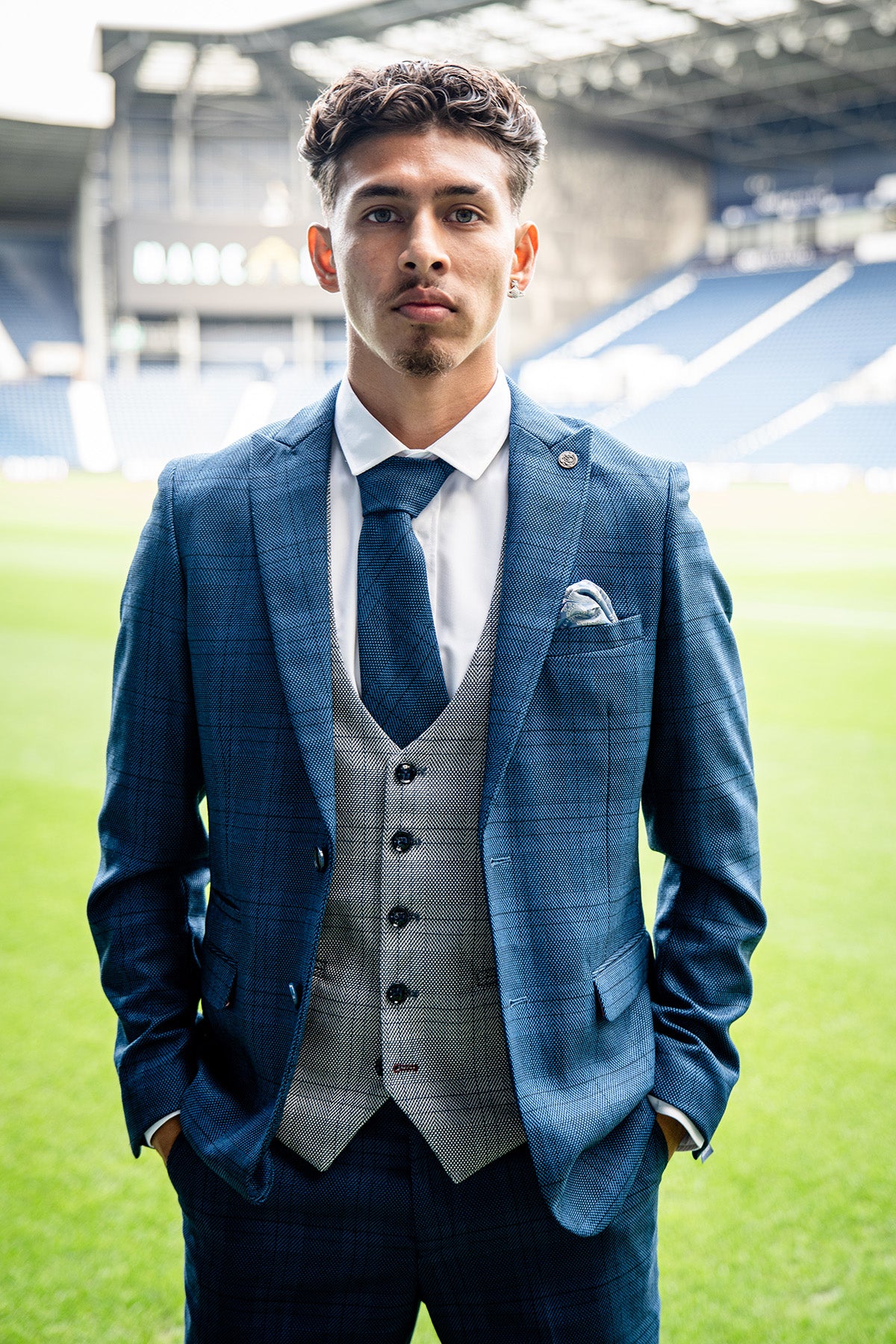 A young man wears the marcdarcy WBA Collection—JERRY Blue Suit with Jerry Grey Waistcoat, as worn by Jeremy Sarmiento, paired with a white shirt and navy tie, standing confidently on a football field with stadium seating behind.