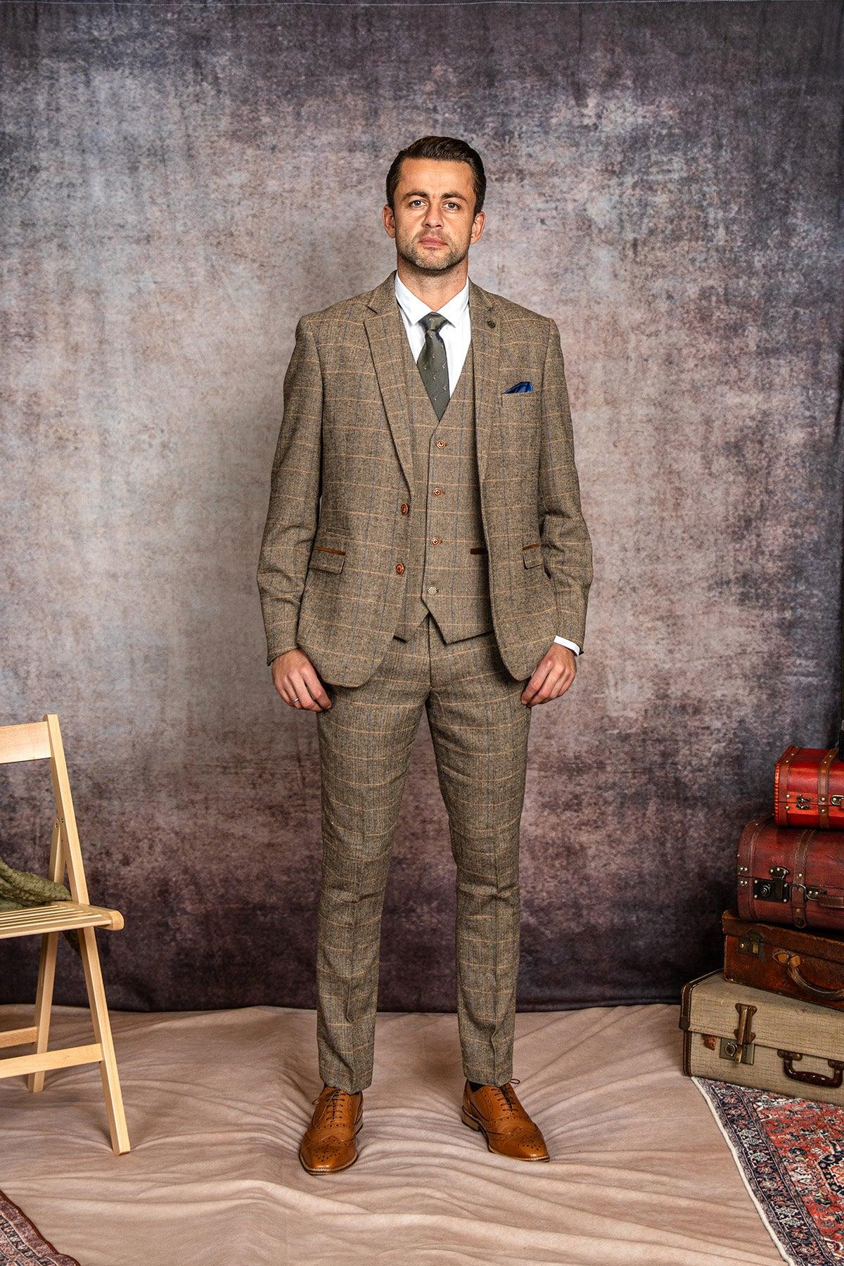 A man stands against a textured backdrop wearing the marcdarcy WHU Collection - TED Tan Tweed Check Three Piece Suit as worn by Lukasz Fabianski, paired with a white shirt, grey tie, and brown dress shoes.
