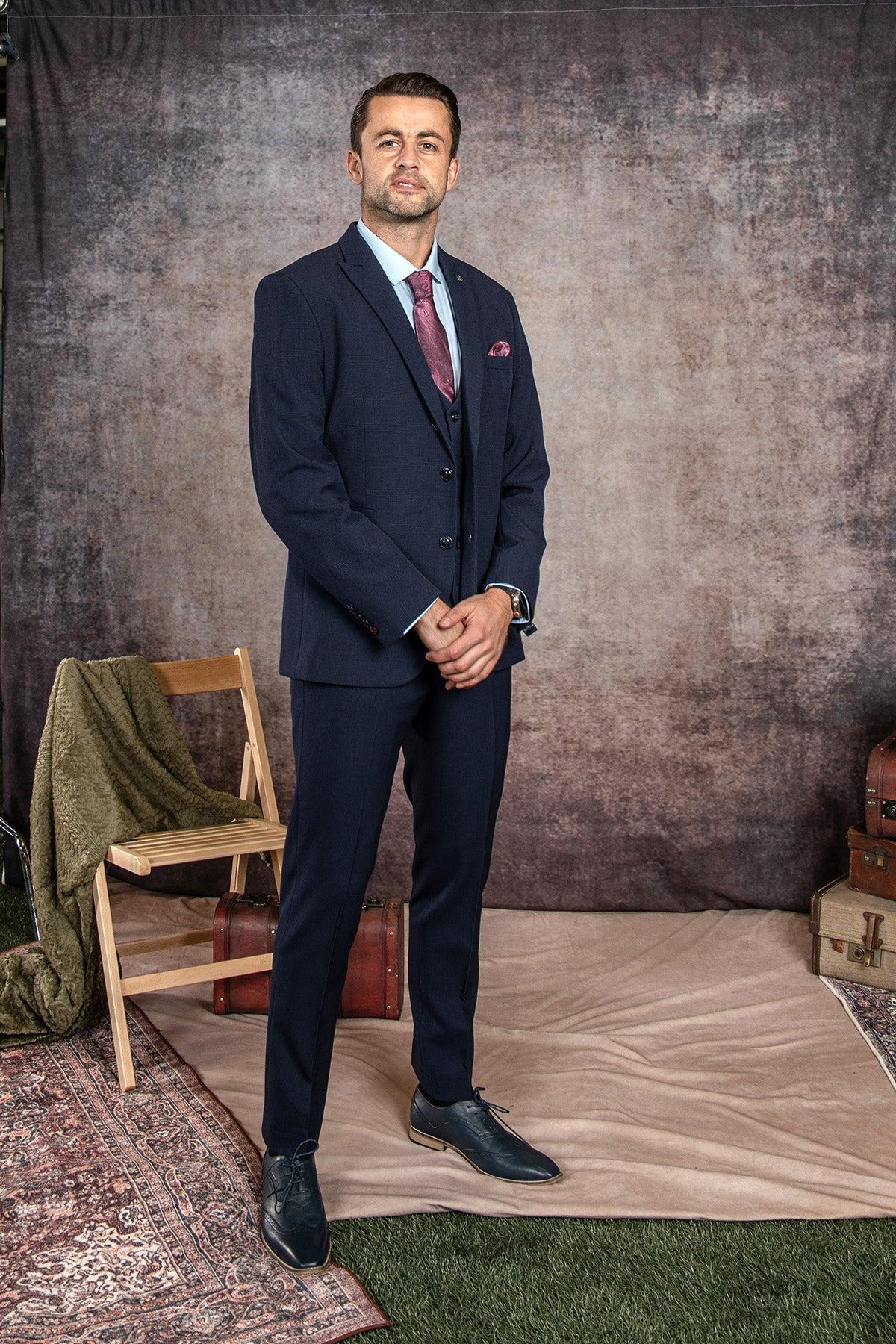 A man wearing the Marc Darcy WHU Collection - BROMLEY Navy Check Three Piece Suit, as worn by Lukasz Fabianski, stands on a beige rug with a wooden chair draped in a green blanket and stacked suitcases behind him.