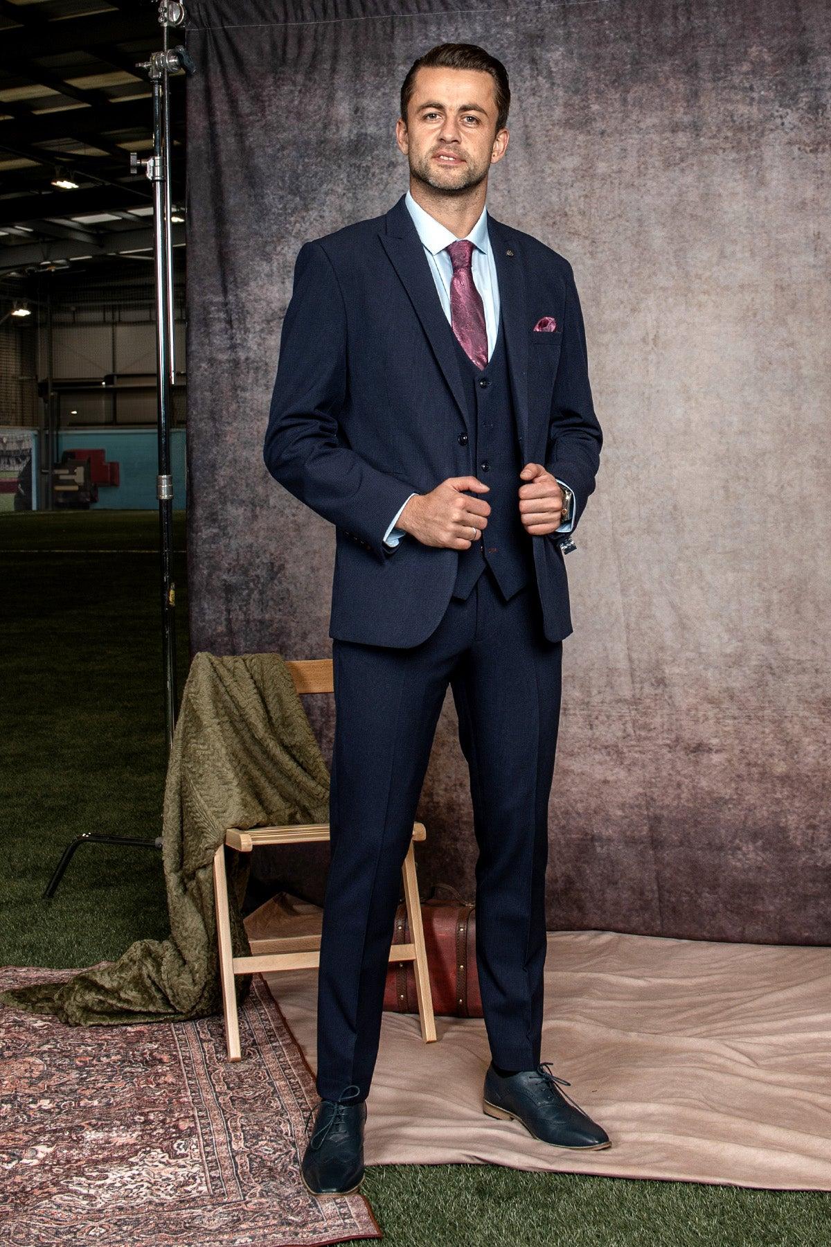 A man wears the Marc Darcy WHU Collection - BROMLEY Navy Check Three Piece Suit as worn by Lukasz Fabianski, styled with a light blue shirt and maroon tie, standing indoors on a rug before a wooden chair and green blanket.