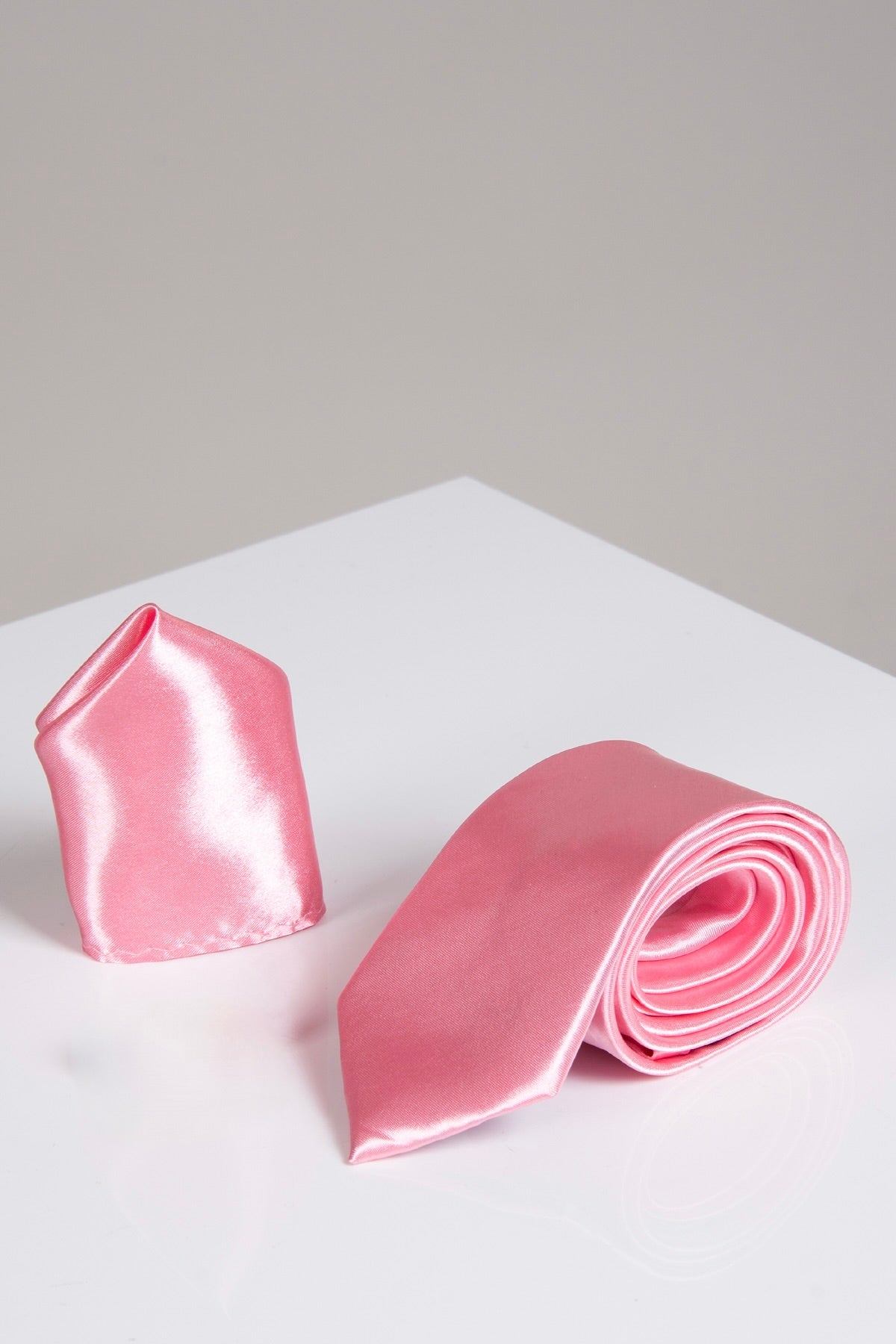 The Accessory ST - Satin Tie and Pocket Square Set in Blush Pink features a shiny satin tie rolled up alongside its matching pocket square, both elegantly displayed on a glossy white surface against a neutral backdrop.