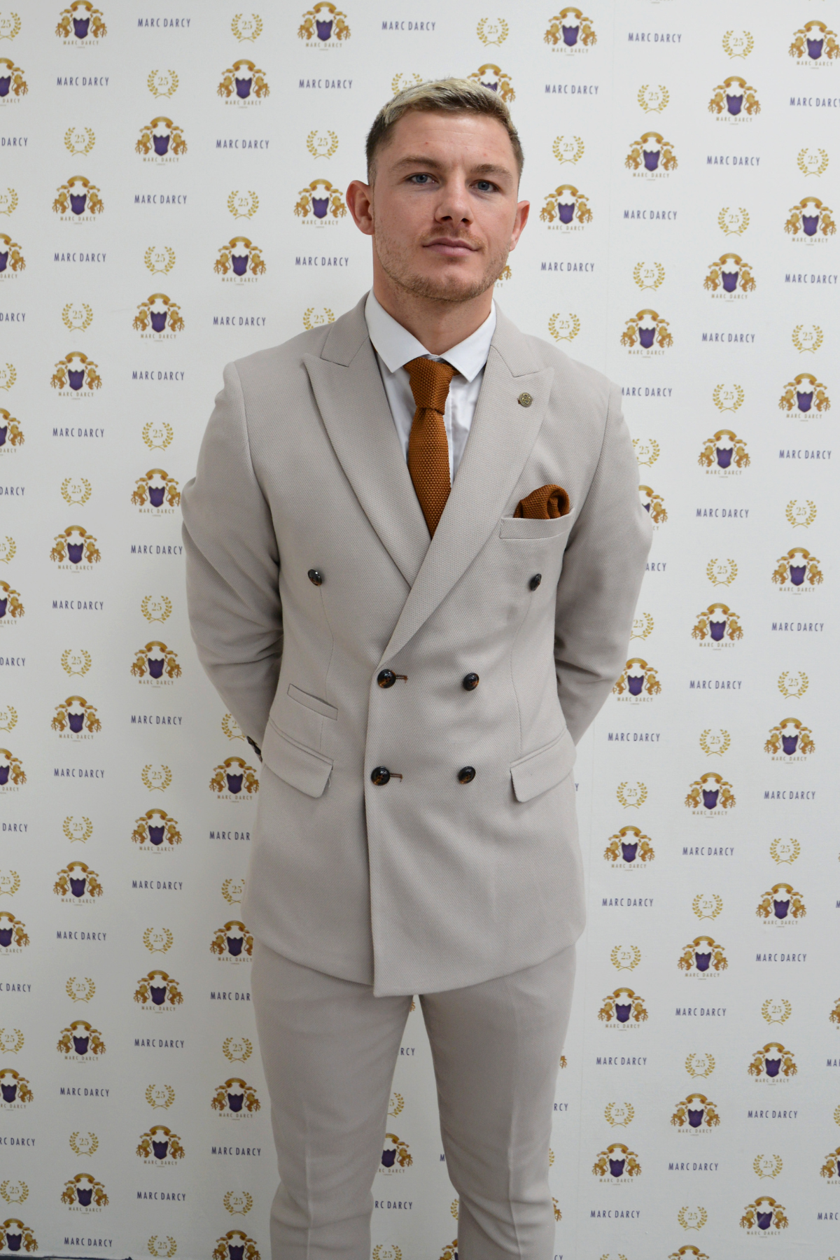 MK Dons footballer Callum Hendry wears a Marc Darcy HM5 Stone double-breasted suit, brown tie, and pocket square while standing before a white backdrop featuring repeating Marc Darcy logos and crests.