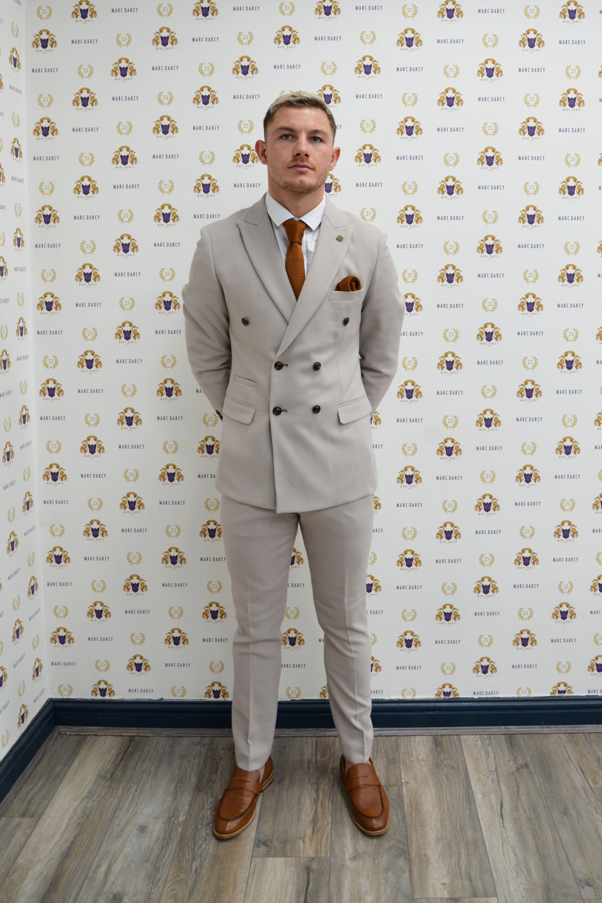 MK Dons footballer Callum Hendry wears a Marc Darcy HM5 Stone double-breasted suit, brown tie, and shoes, standing on a wooden floor before a repeating circular Rate Limit logo backdrop with his hands behind his back.
