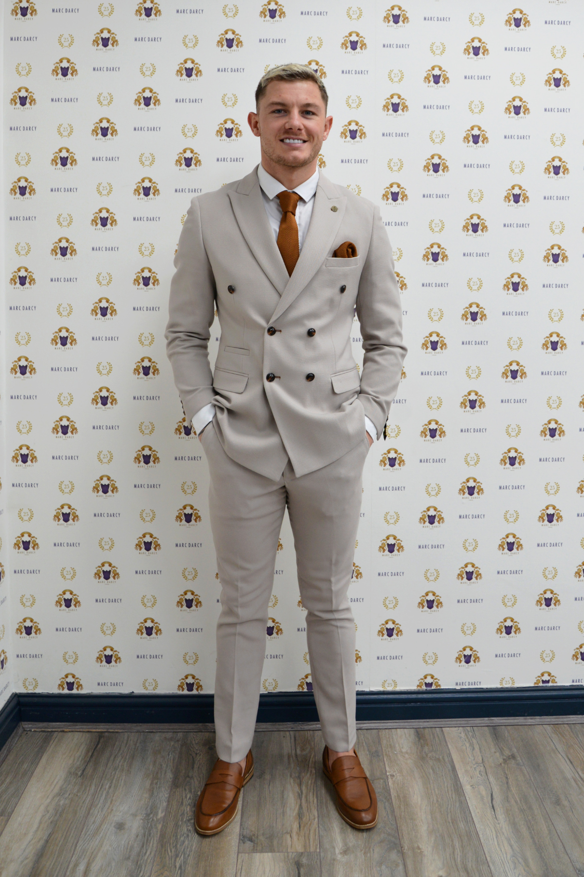 MK Dons footballer Callum Hendry wears the Marc Darcy HM5 Stone Double-Breasted Suit, paired with a white shirt and orange tie, standing against a wall with a repeated Marc Darcy logo pattern.