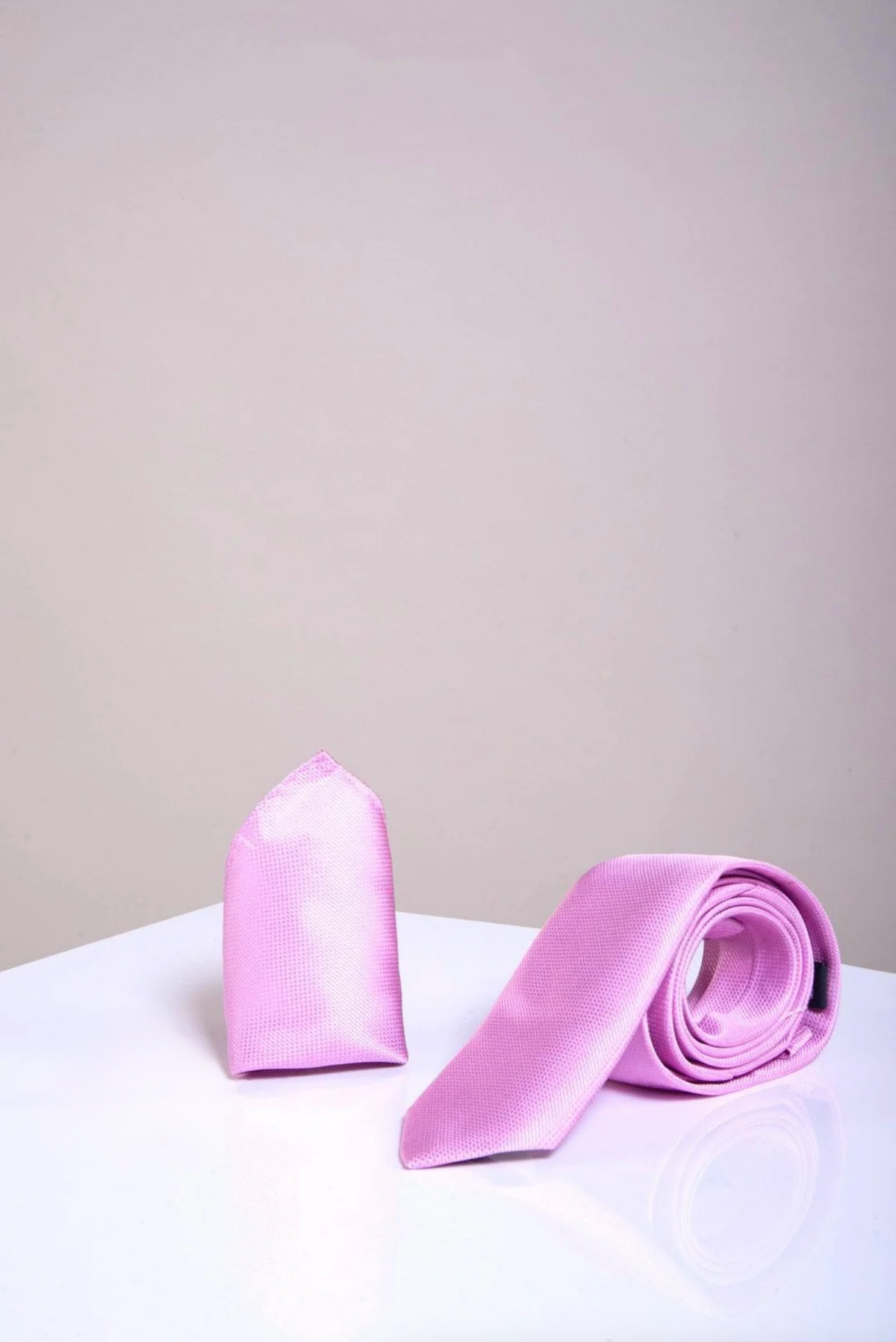 The Accessory TB17 Birdseye Tie and Pocket Square Set in Pink features a folded pocket square and a rolled necktie, both displayed upright on a glossy white surface against a plain light gray background.