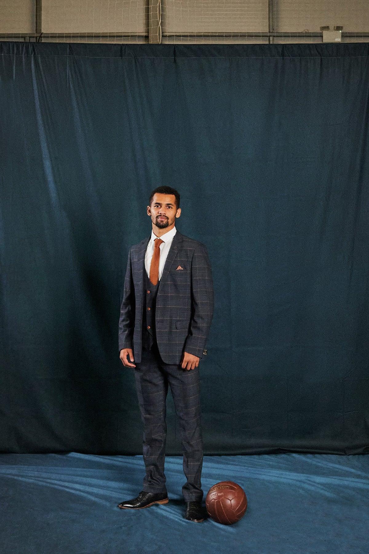 A man wears the marcdarcy JENSON Marine Navy Check Suit from The Everton Collection, white shirt, and tie, standing on a blue floor near a brown vintage basketball with a dark blue curtain backdrop.