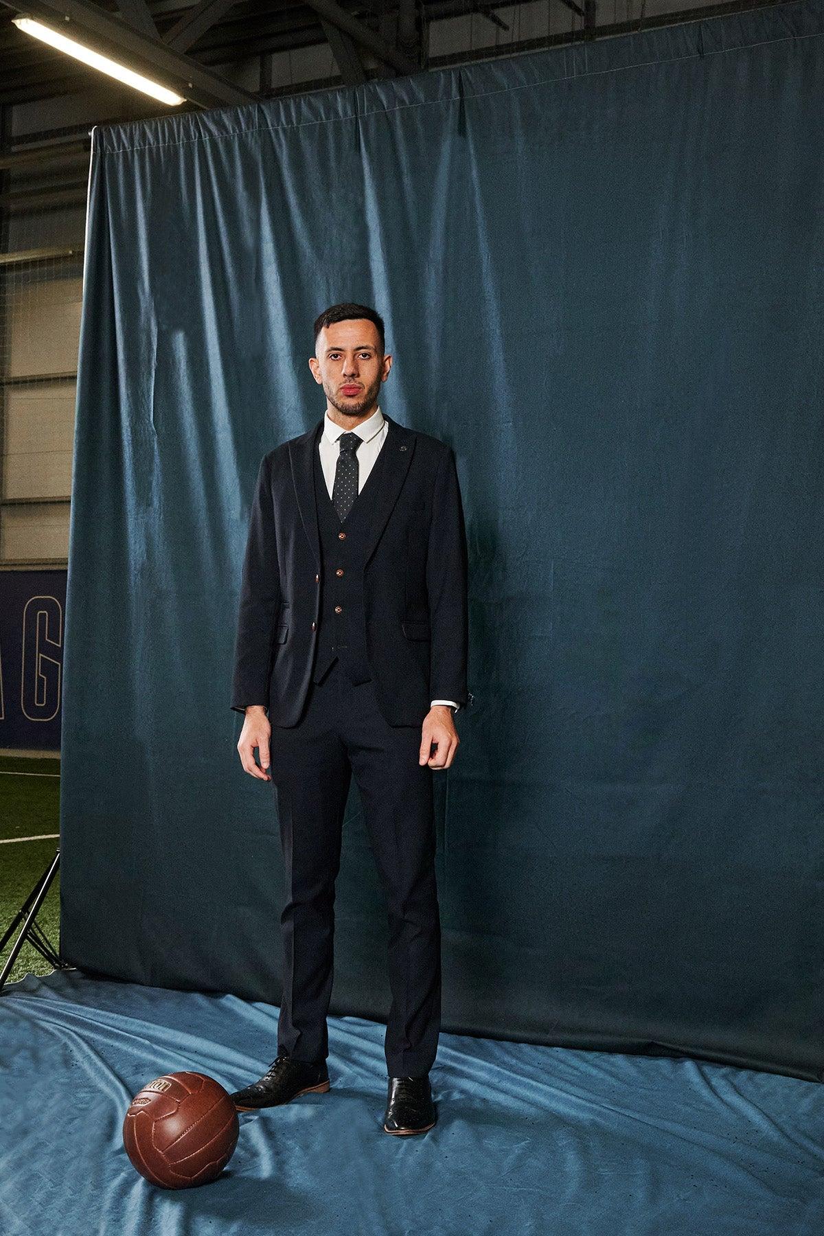 A man wears the marcdarcy Everton Collection Dwight McNeil in Max Navy Suit and tie, standing indoors on blue fabric by a vintage brown soccer ball, with a blue curtain backdrop evoking sports-facility style.