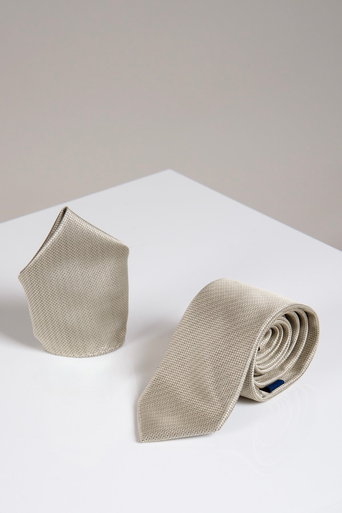 The Accessory TB17 Birdseye Tie and Pocket Square Set in Tan, featuring a textured necktie and matching pocket square, is shown on a glossy white surface against a light gray background.