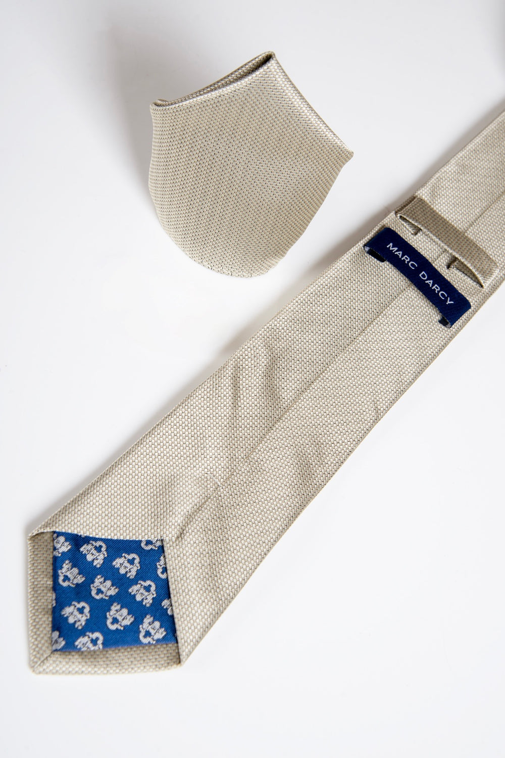 The TB17 - Birdseye Tie and Pocket Square Set in Tan by Accessory is displayed on a white surface. The tie is turned over, showing its blue floral lining and a label with the brand name "Accessory.
