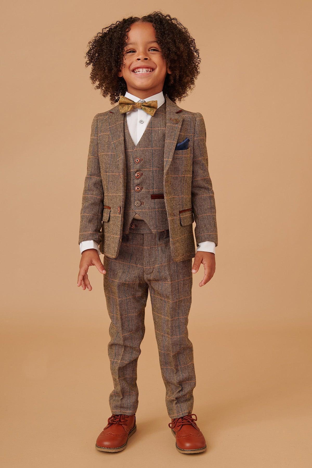 A young child with curly hair smiles against a beige background, wearing the marcdarcy TED Childrens Tan Tweed Check Three Piece Suit, featuring a matching vest, white shirt, gold bow tie, and brown shoes.