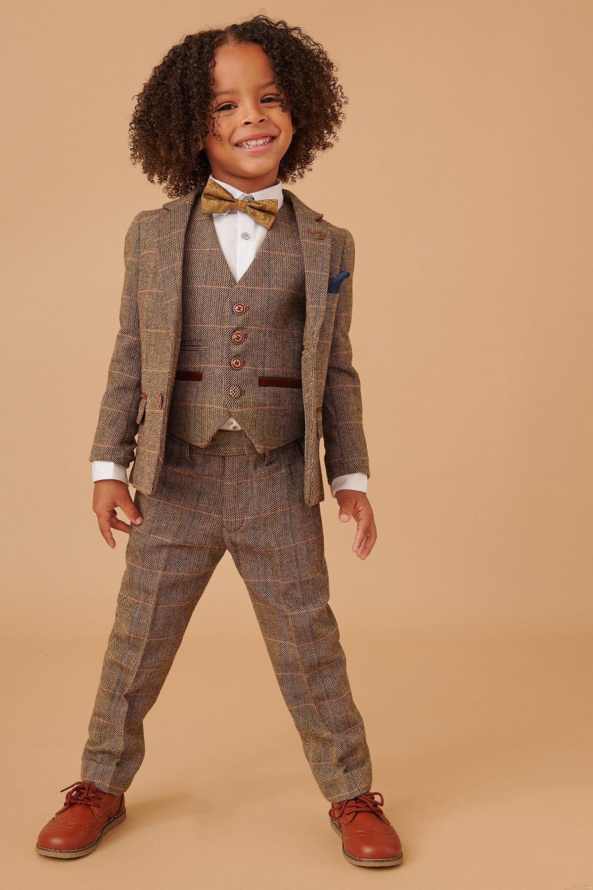 A young child smiles in the marcdarcy TED Children's Tan Tweed Check Three Piece Suit—ideal as a kids' wedding outfit for a country barn setting—styled with a white shirt, yellow bow tie, and brown shoes against a plain beige backdrop.