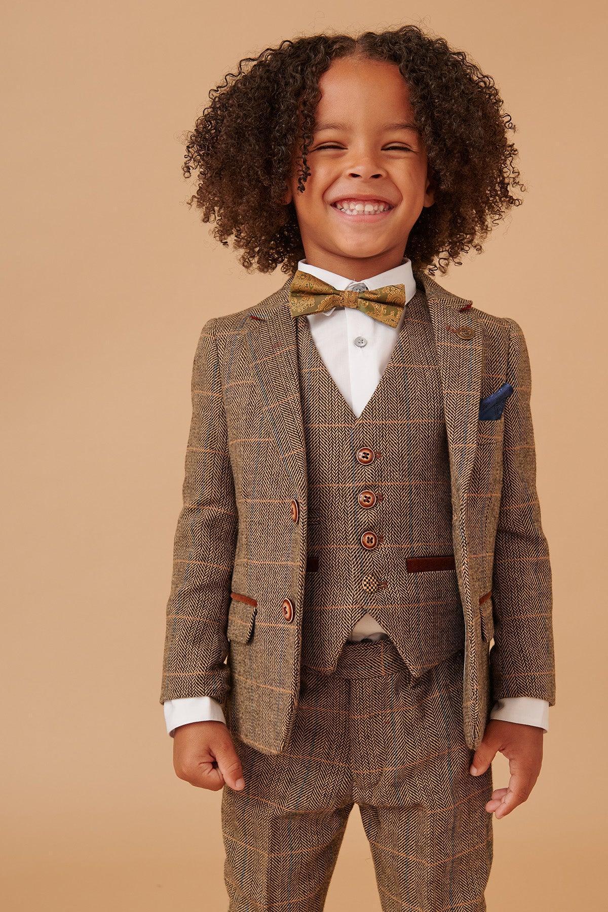 A young child with curly hair smiles widely while wearing the marcdarcy TED Childrens Tan Tweed Check Three Piece Suit with a matching vest, bow tie, and crisp white shirt, standing against a beige background.