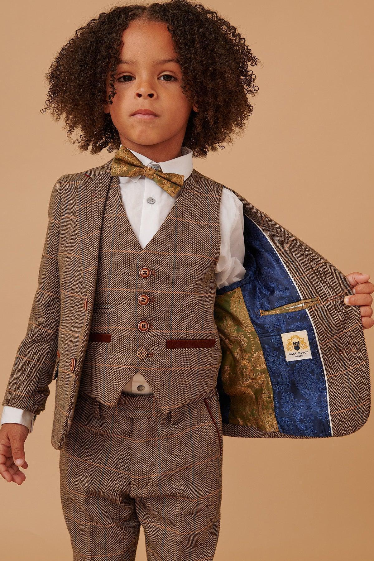 A young boy with curly hair models the marcdarcy TED Childrens Tan Tweed Check Three Piece Suit, including a vest, white shirt, and bow tie. He confidently reveals the suit’s patterned blue and green lining against a beige background.