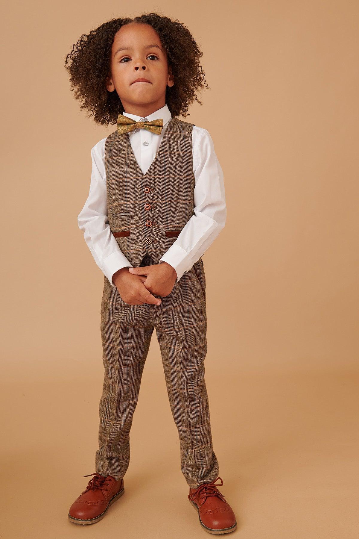 A young boy stands confidently against a beige background, dressed in the marcdarcy TED Childrens Tan Tweed Check Three Piece Suit featuring a stylish plaid vest and pants, white shirt, gold bow tie, and brown dress shoes.