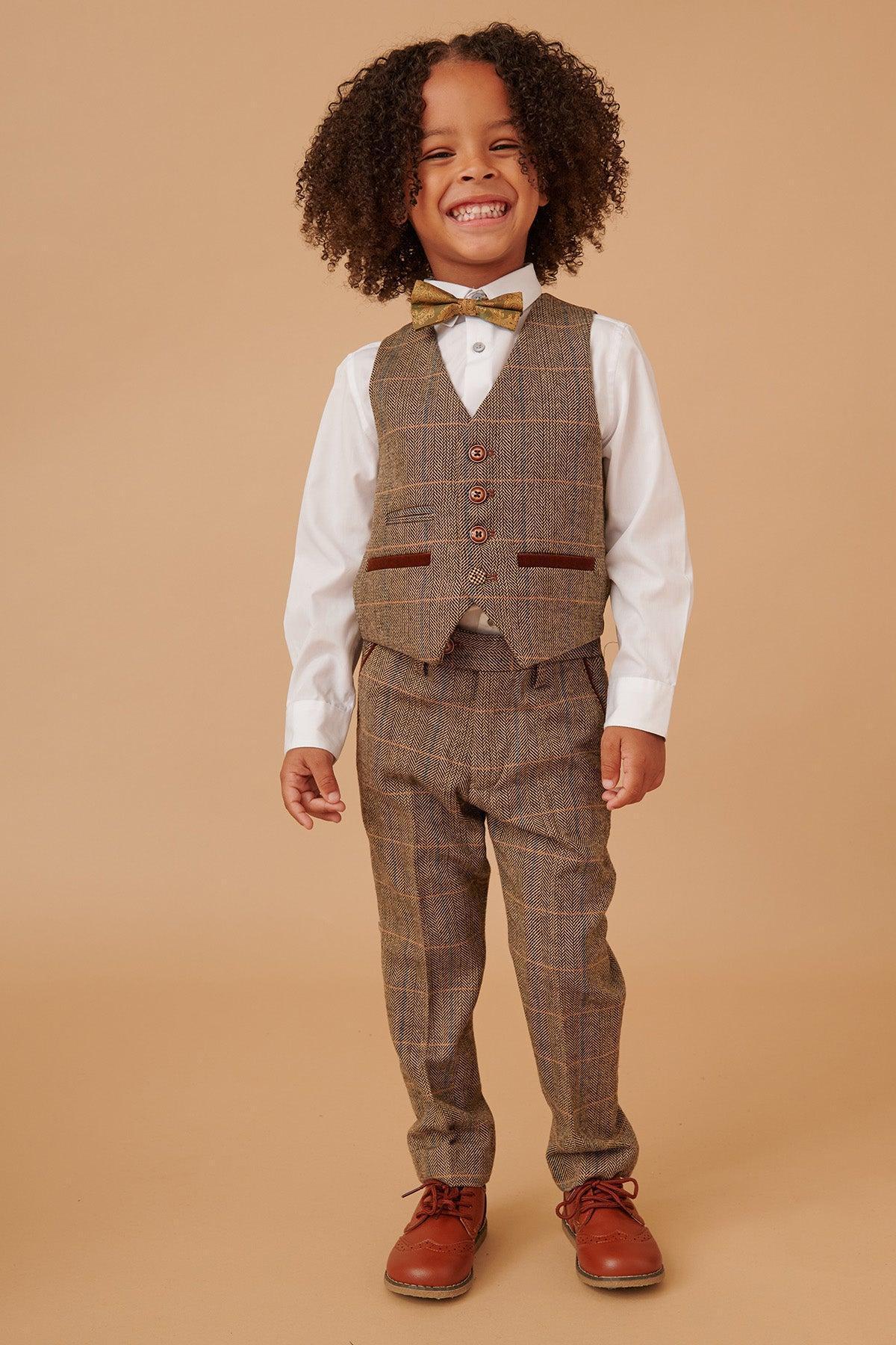 Smiling young boy with curly hair wears the marcdarcy TED Childrens Tan Tweed Check Three Piece Suit, featuring a vest and pants, white shirt, gold bow tie, and brown shoes, standing against a plain beige background.