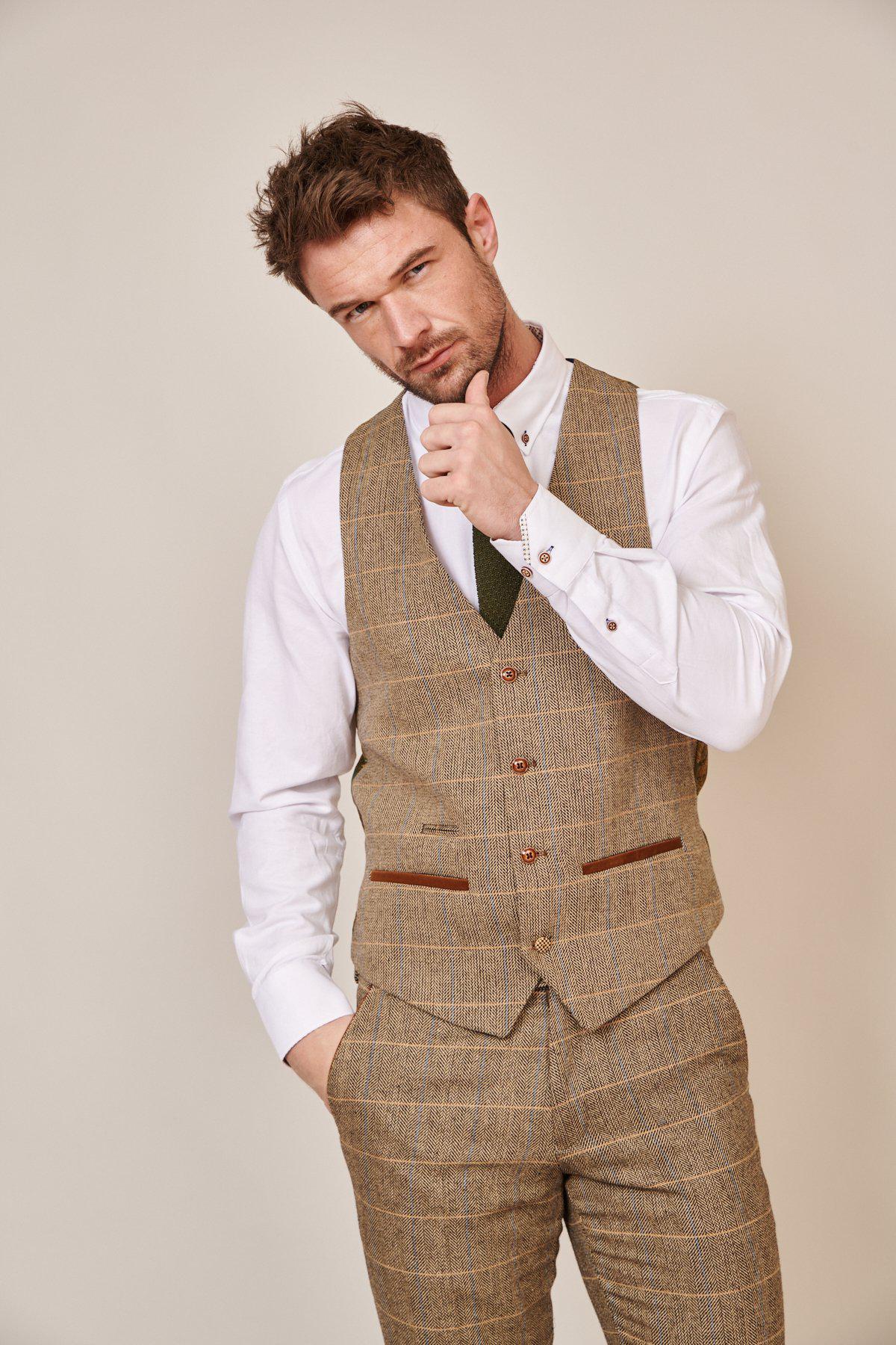 A man in the marcdarcy WHU Collection - TED Tan Tweed Check Three Piece Suit, as worn by Lukasz Fabianski, stands against a plain background, wearing a white shirt and striking a thoughtful pose with one hand on his chin.