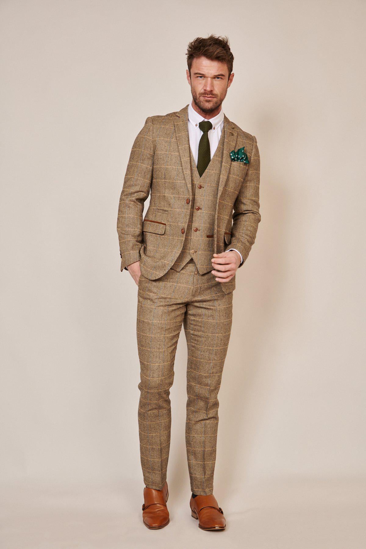 A man wears the marcdarcy WHU Collection - TED Tan Tweed Suit with Dion Waistcoat as worn by Lukasz Fabianski, paired with a white shirt, olive green tie, pocket square, and brown shoes. He has short hair, a trimmed beard, and poses one hand in pocket.
