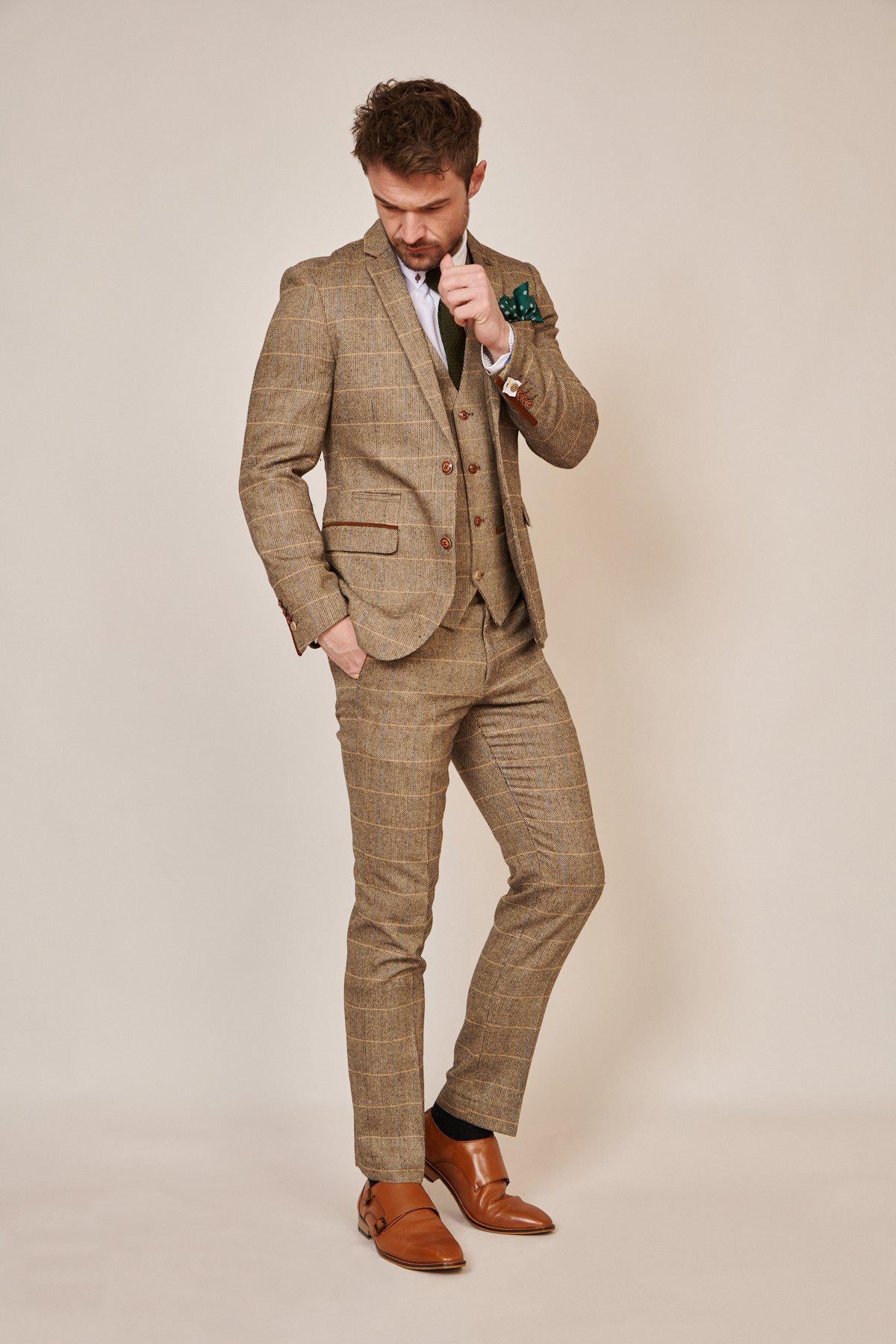 A man stands against a plain background in marcdarcy’s WHU Collection - TED Tan Tweed Check Three Piece Suit, as worn by Lukasz Fabianski, with a white shirt, green pocket square, and tan dress shoes, adjusting his tie in timeless English style.