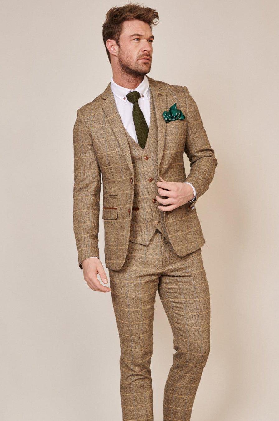 A man models marcdarcy’s WHU Collection – TED Tan Tweed Suit with Dion Waistcoat, as worn by Lukasz Fabianski, paired with a white shirt, olive green tie, and green polka dot pocket square against a beige backdrop.