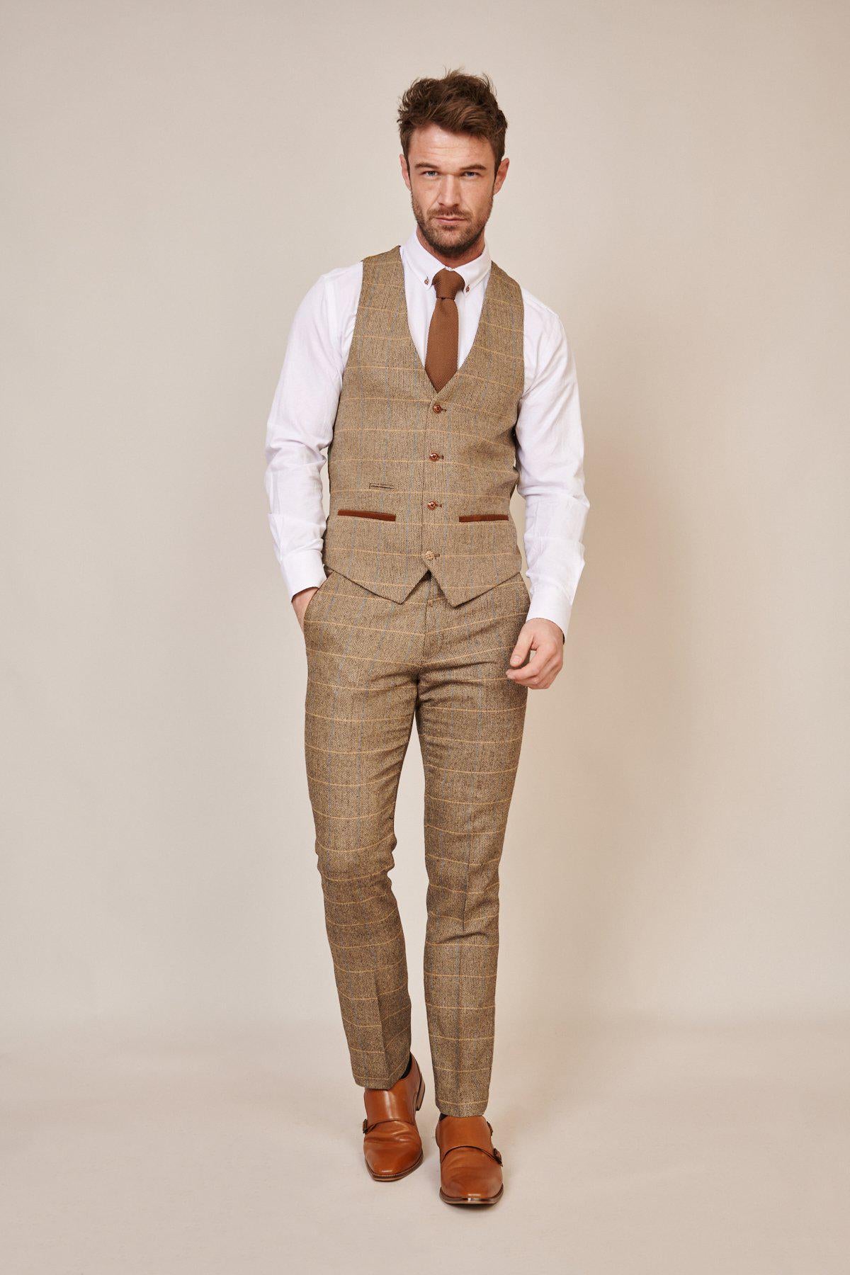 A man wearing the marcdarcy WHU Collection - TED Tan Tweed Check Three Piece Suit as worn by Lukasz Fabianski stands against a plain beige background, pairing it with a white dress shirt, brown tie, and brown shoes, one hand in his pocket.
