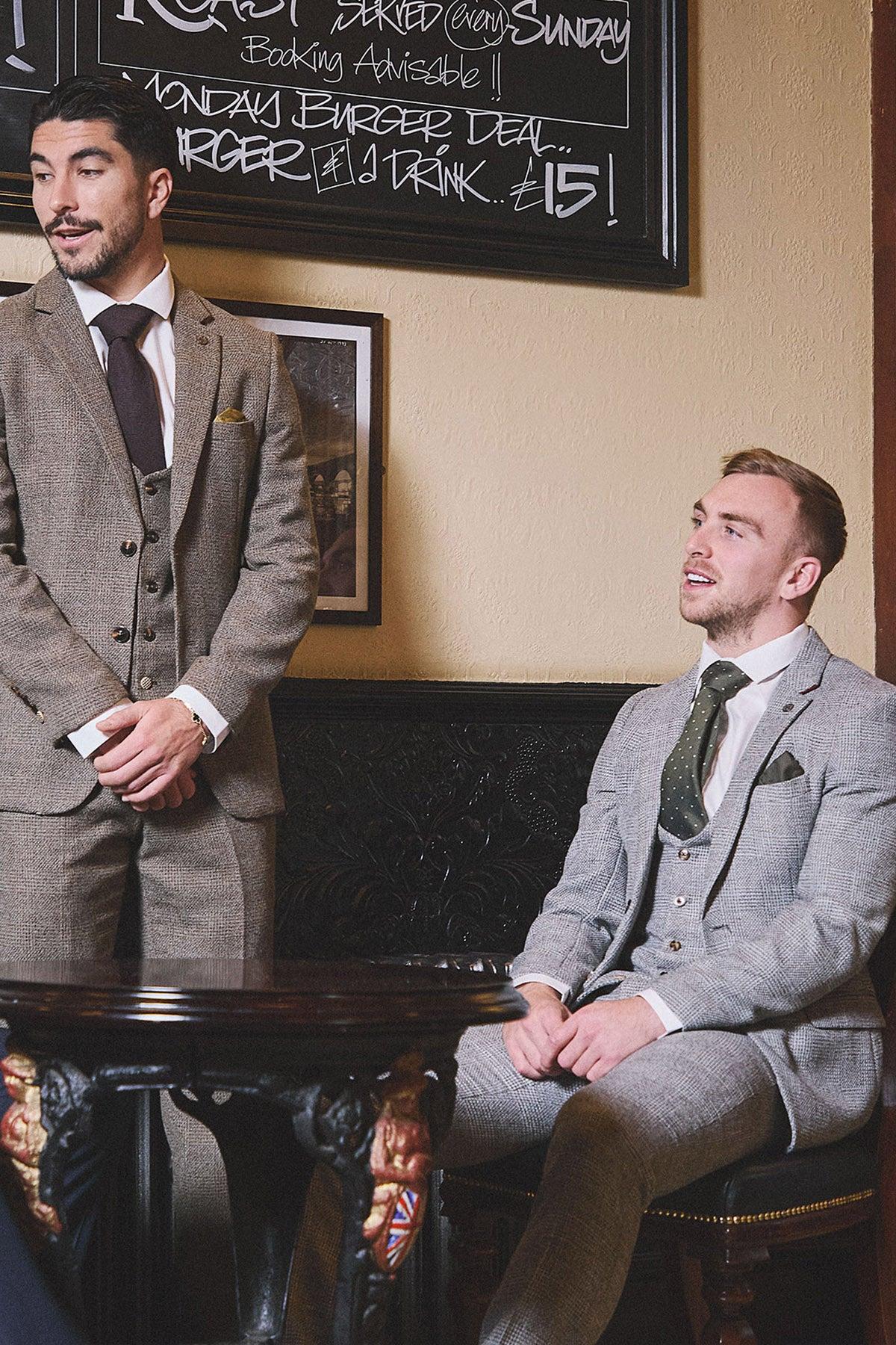 Two men in marcdarcy’s WHU Collection - KRIS Silver Grey Tweed Suits, as worn by Jarrod Bowen, sit at a table with a chalkboard menu behind them.