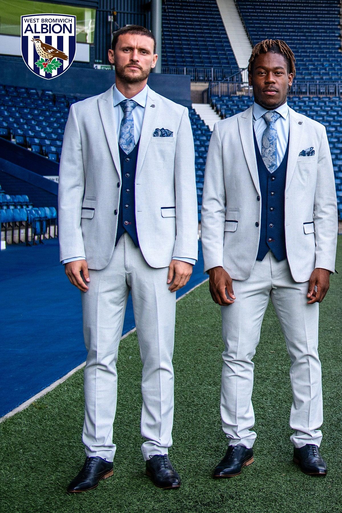 Two men in Marc Darcys WBA Collection BROMLEY Stone Suit with Kelvin Navy Waistcoat and patterned ties stand seriously on a football pitch, with stadium seats and the West Bromwich Albion logo visible in the background.