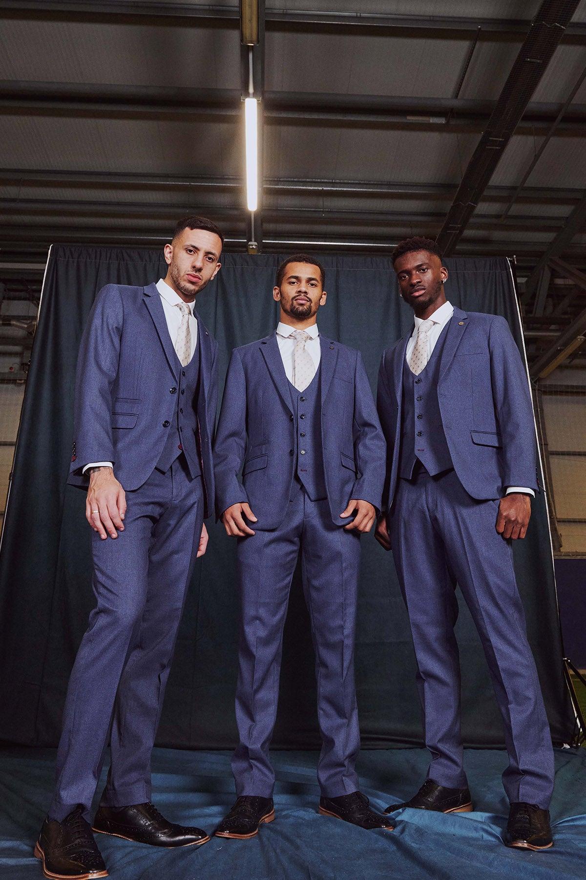 Three men in matching Marc Darcy’s The Everton Collection | ASHLEY Navy Blue Three Piece Suits, white shirts, and patterned ties stand confidently before a dark backdrop, facing the camera.