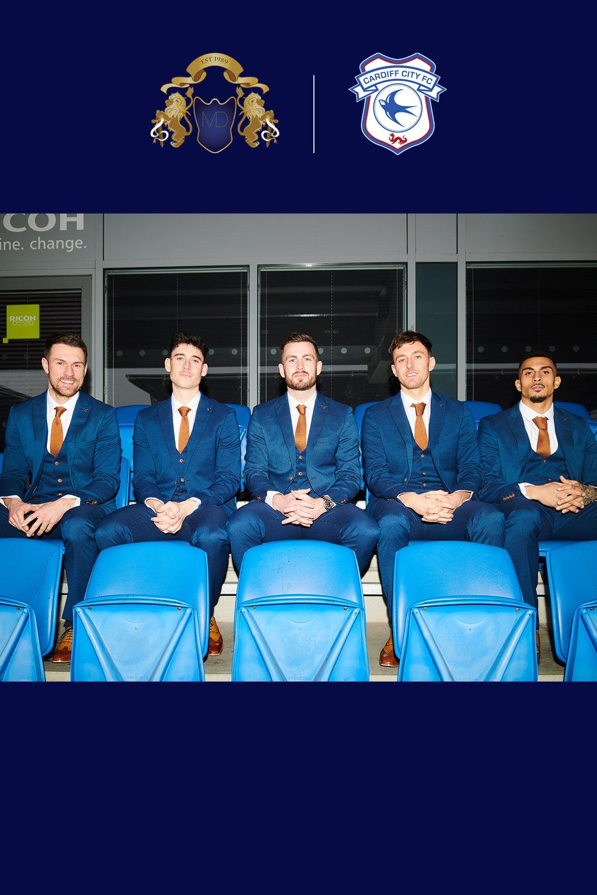 Five men wear marcdarcy Official Cardiff City F.C. Club Suits (MAX Royal Blue) with orange ties, sitting in blue stadium seats and smiling. Two logos, featuring gold lions and Cardiff City FC, appear above on a navy background.