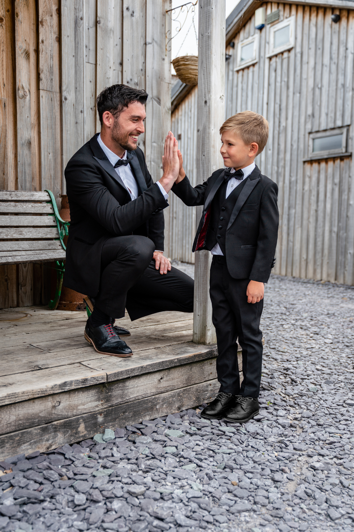 A man and a boy smile and high-five on a wooden porch, both wearing marcdarcy’s Matching Father & Son DALTON Black Tux Three Piece Suits.