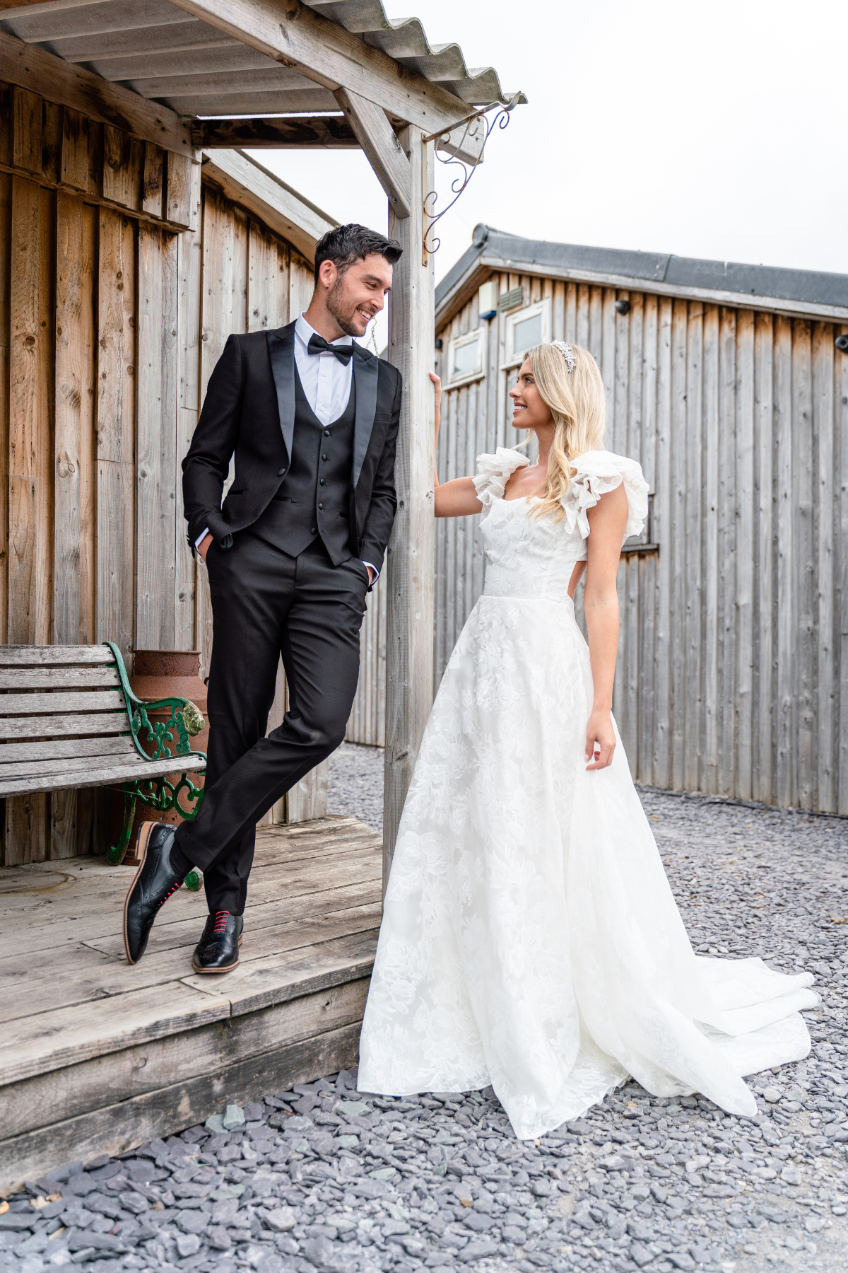 A groom in the marcdarcy Matching Father & Son | DALTON Black Tux Three Piece Suit smiles at his bride on a wooden porch, rustic wooden buildings behind them.