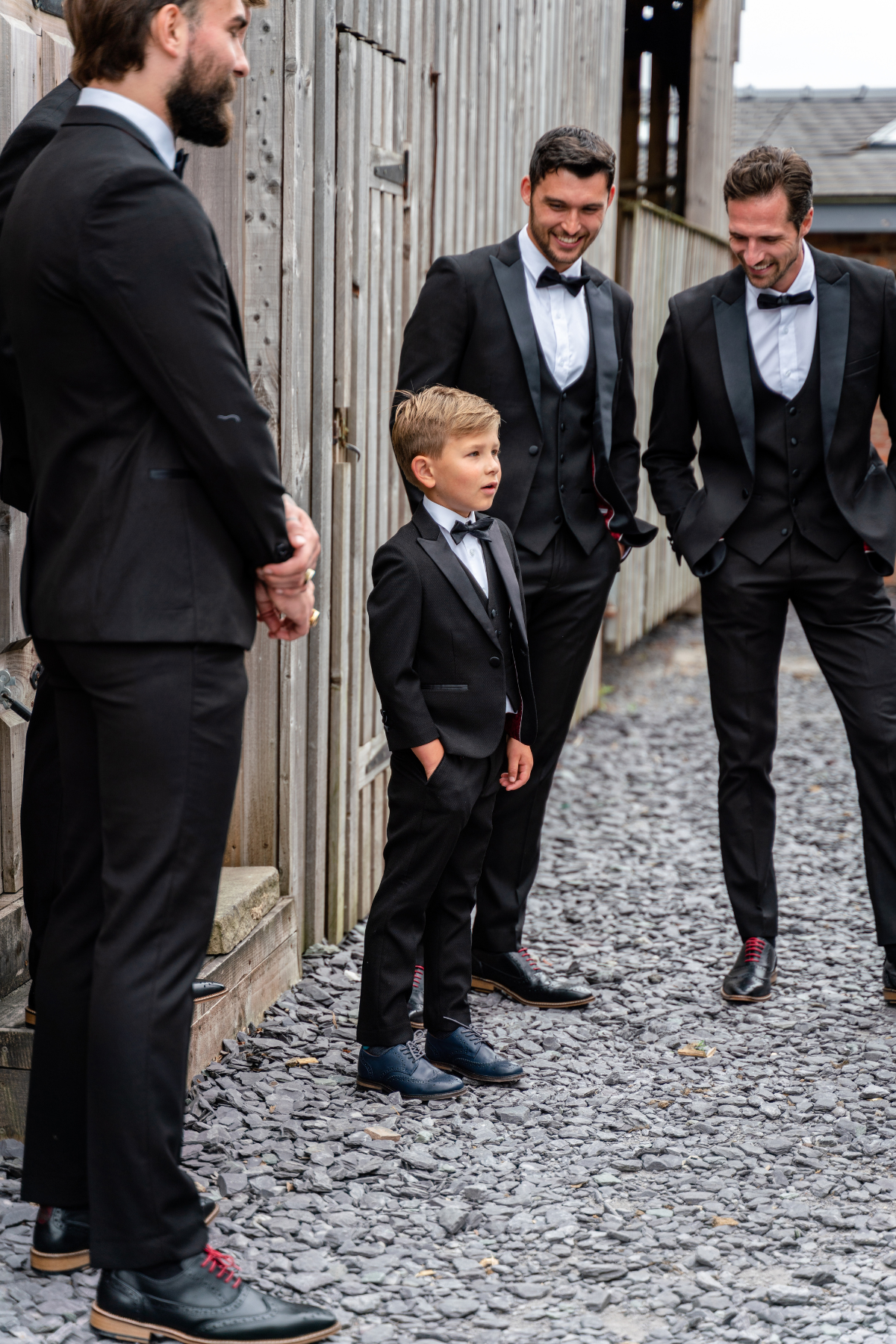 A young boy in a DALTON - Black Tux Three Piece Suit by marcdarcy stands with his hands in his pockets, looking surprised, next to three men wearing matching marcdarcy tuxedos.