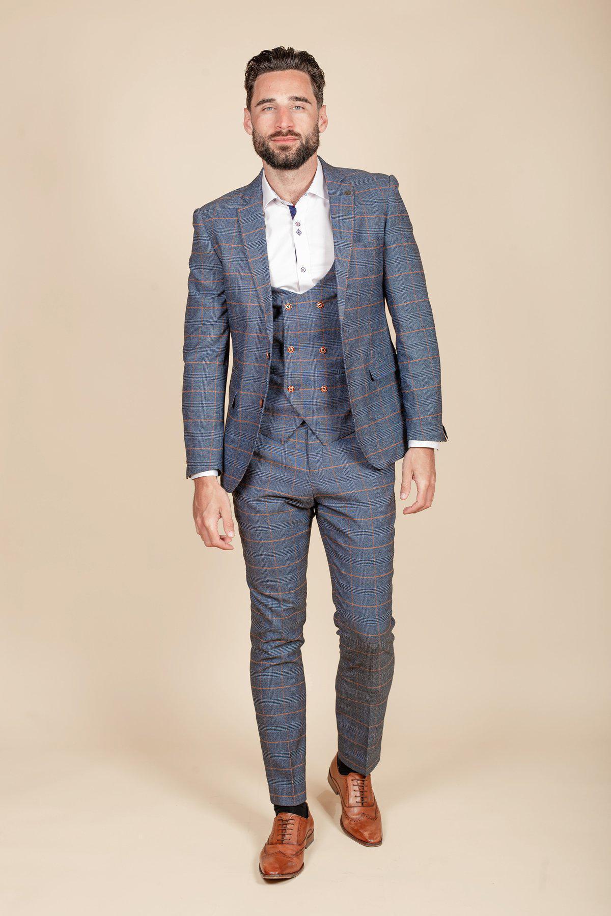 Daniel Osbourne (Rob Mallard) from Coronation Street wears the marcdarcy JENSON Sky Blue Check Suit with a white shirt and brown dress shoes, standing against a beige background and smiling slightly.