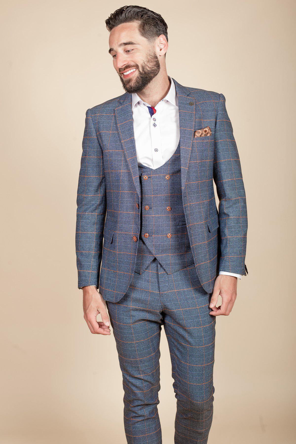Rob Mallard as Coronation Street’s Daniel Osbourne smiles to the side in a marcdarcy JENSON Sky Blue Check Suit, white shirt, and brown pocket square, standing against a beige background.