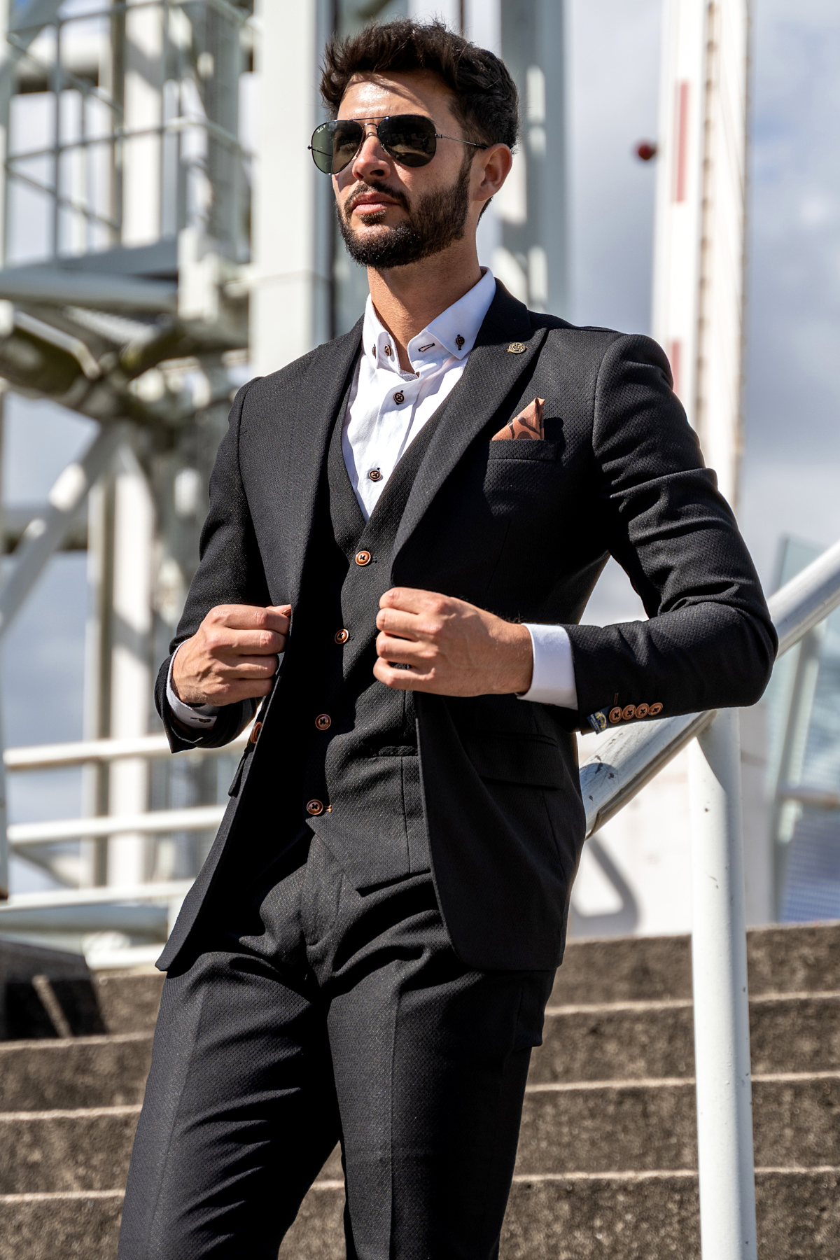 A man in the marcdarcy MAX Black Three Piece Suit with Contrast Buttons and a white shirt adjusts his jacket on concrete steps, with a metal structure and cloudy sky behind him, embodying modern workwear style.