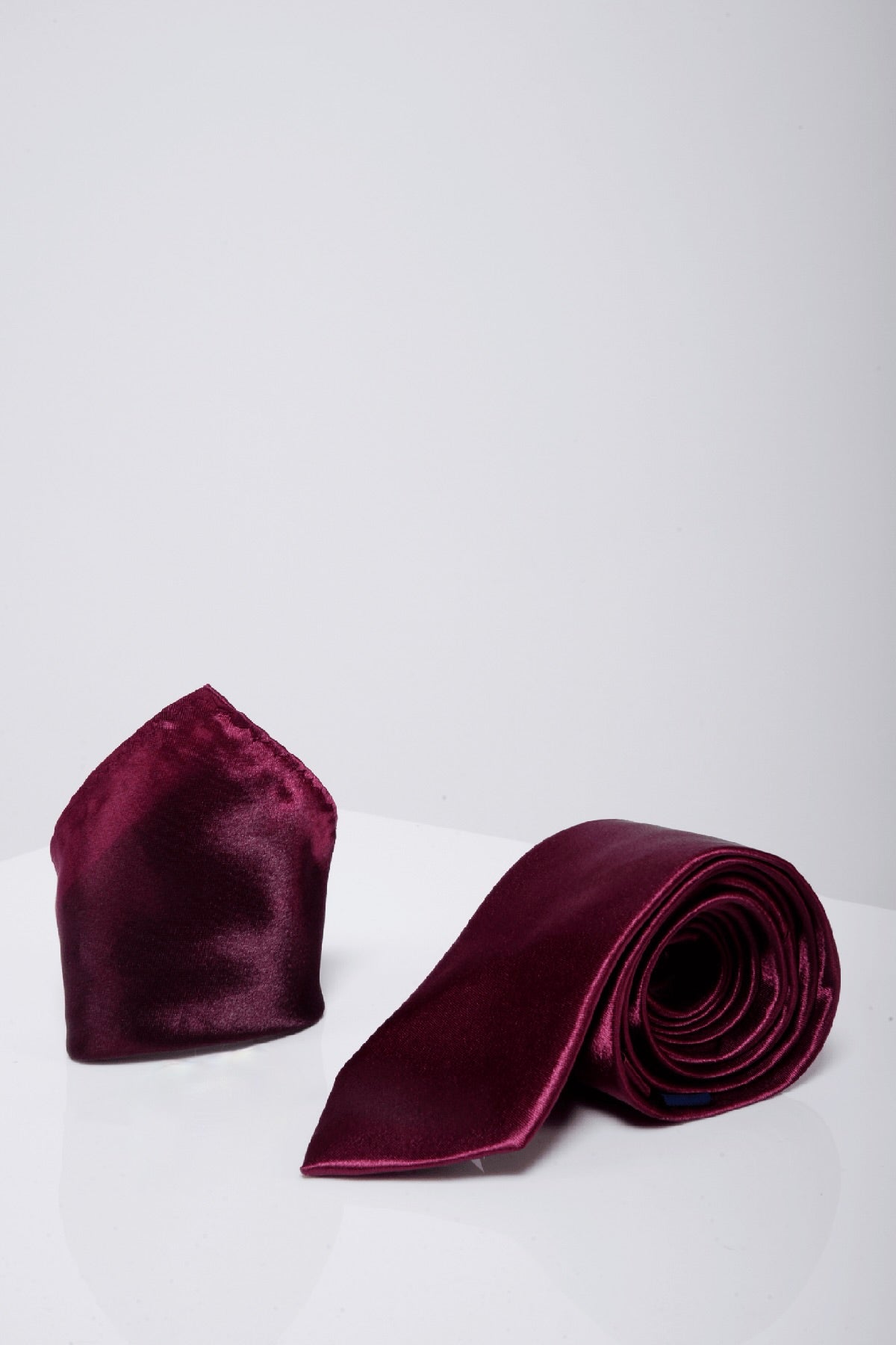 The ST - Satin Tie and Pocket Square Set in Plum by Accessory is displayed on a glossy white surface against a plain light gray background.