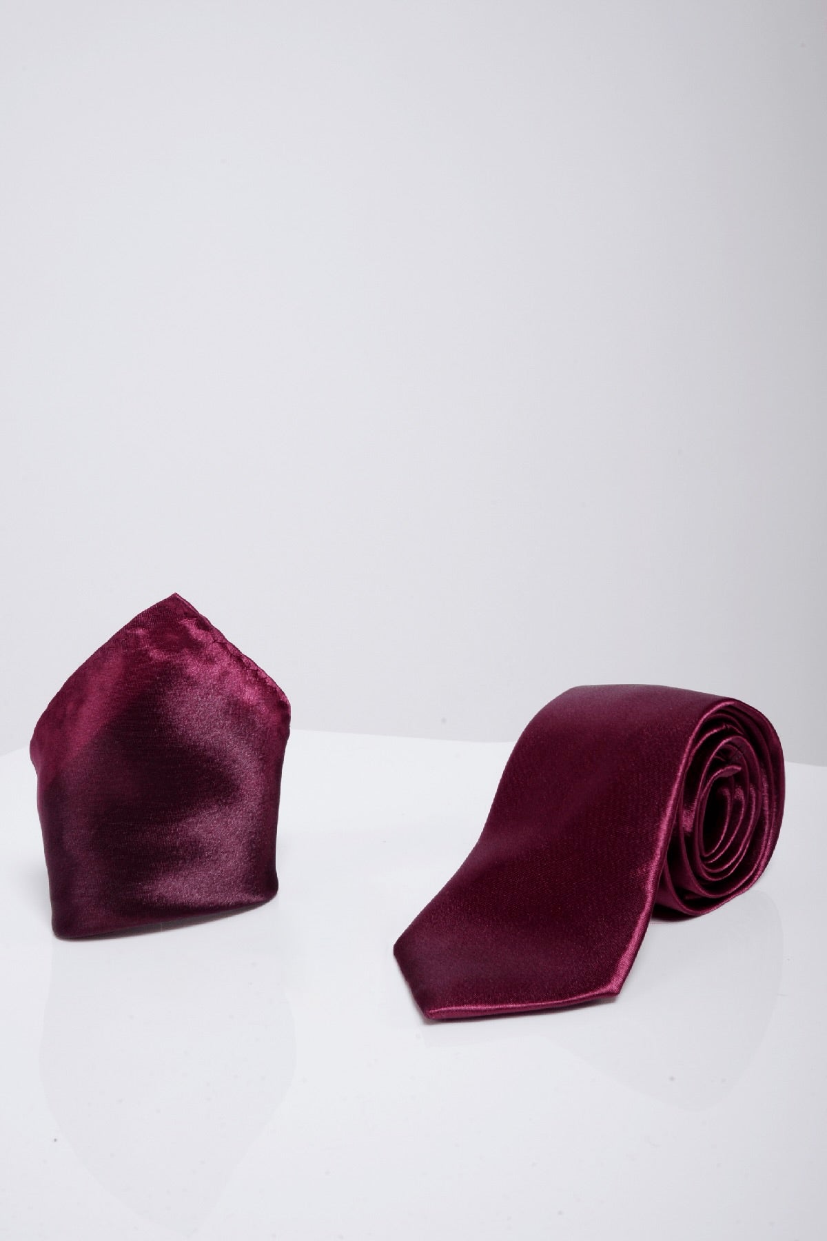 The Accessory ST - Satin Tie and Pocket Square Set In Plum is displayed on a glossy white surface with a plain light gray background, highlighting its elegant and formal style.