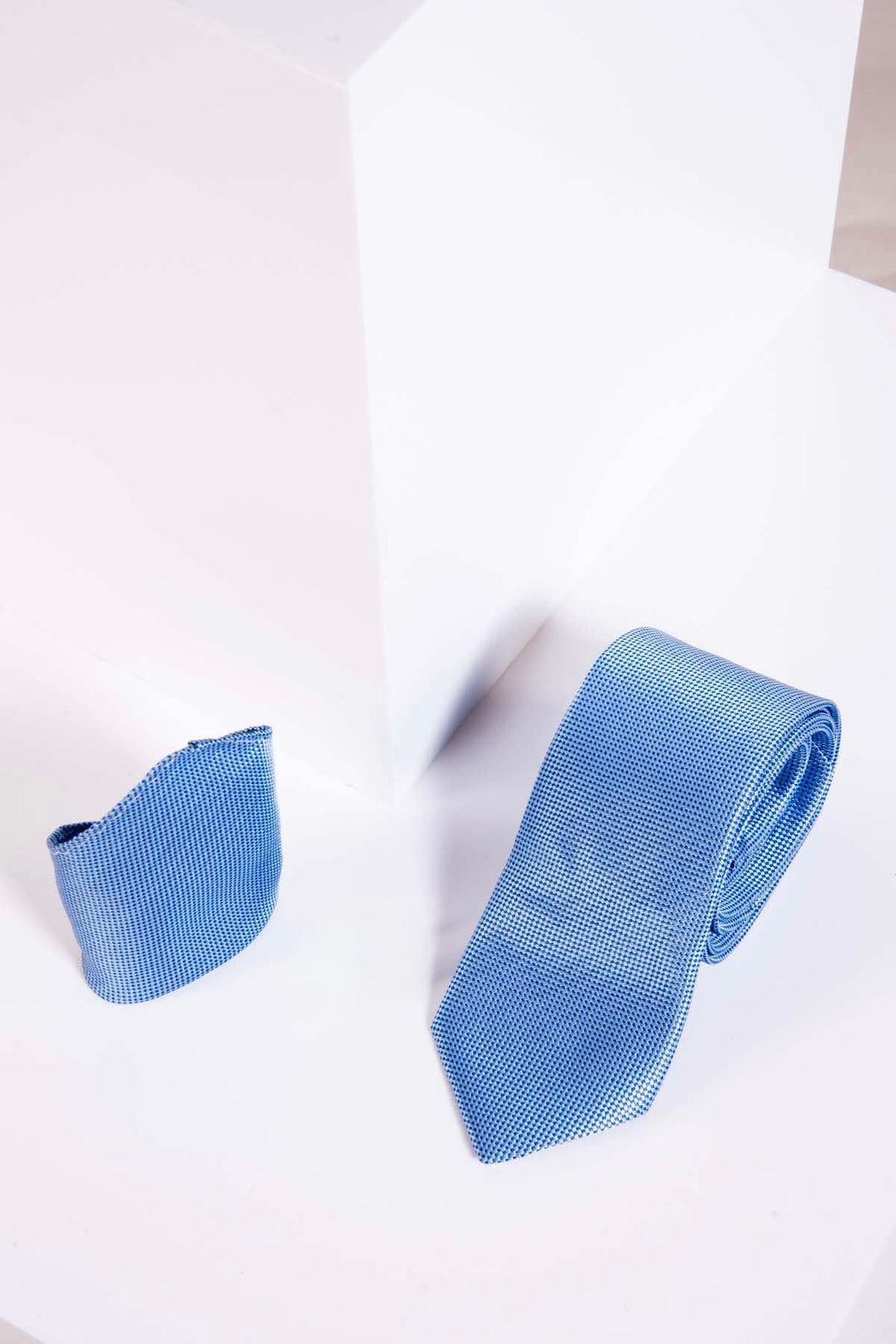 The Accessory TB17 Birdseye Tie and Pocket Square Set in Blue is displayed on a white geometric surface with a white block in the background.