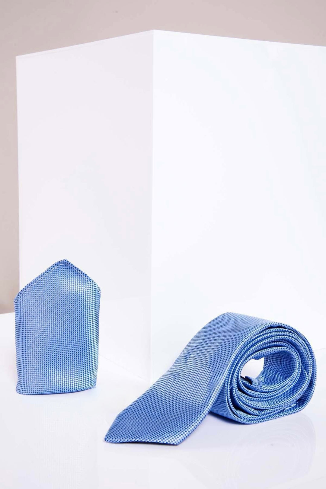 The TB17 - Birdseye Tie and Pocket Square Set in Blue by Accessory is displayed on a glossy white surface before a white cube, offering a simple, modern backdrop.