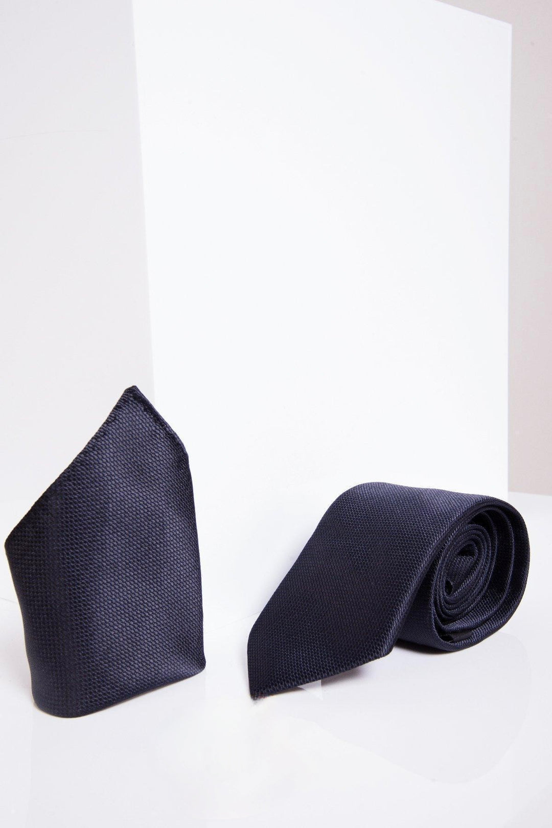 The Marc Darcy TB17 Birdseye Tie and Pocket Square Set in Midnight features a neatly folded pocket square and matching rolled tie, making an ideal wedding accessory—elegantly displayed against a white geometric backdrop.