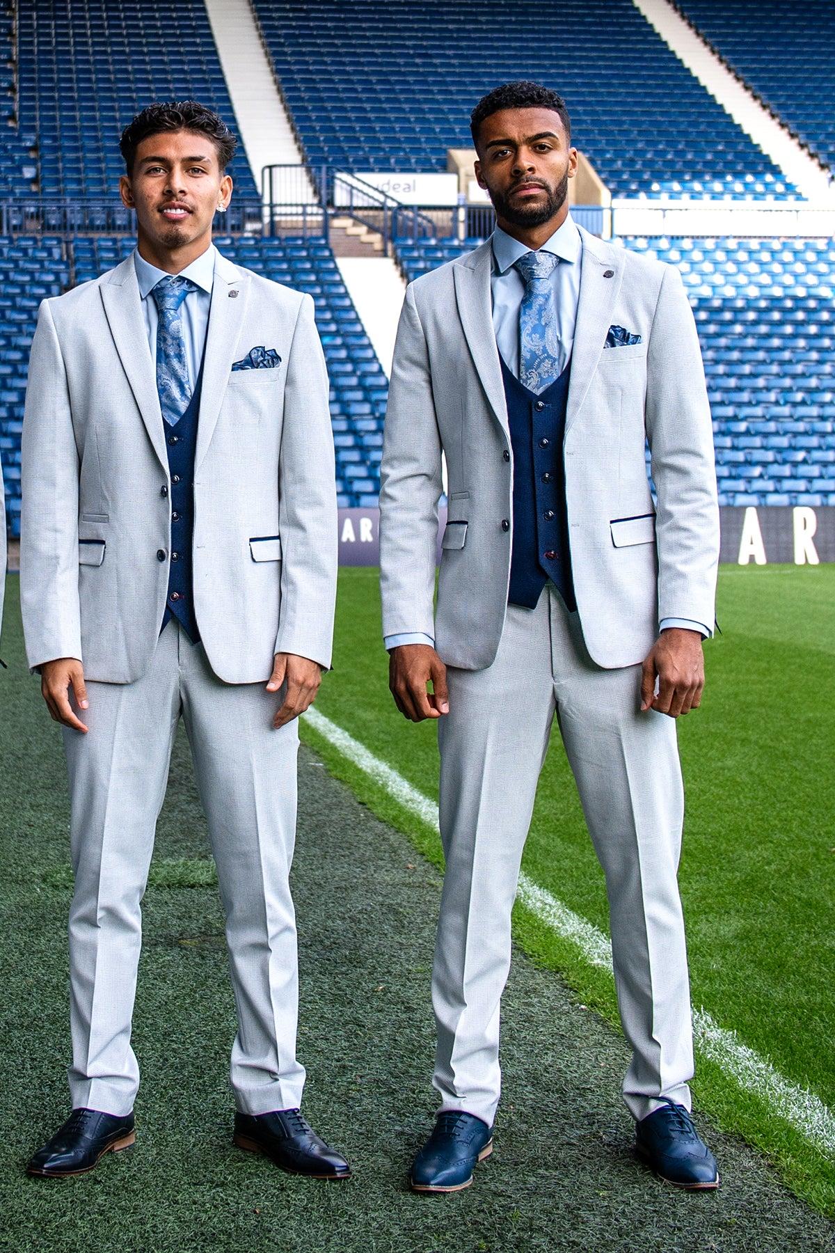 Two men stand on a football field wearing Marc Darcy’s The WBA Collection - BROMLEY Stone Suit with Kelvin Navy Waistcoat, patterned ties, and pocket squares, complementing the stadium seats and field in the background.