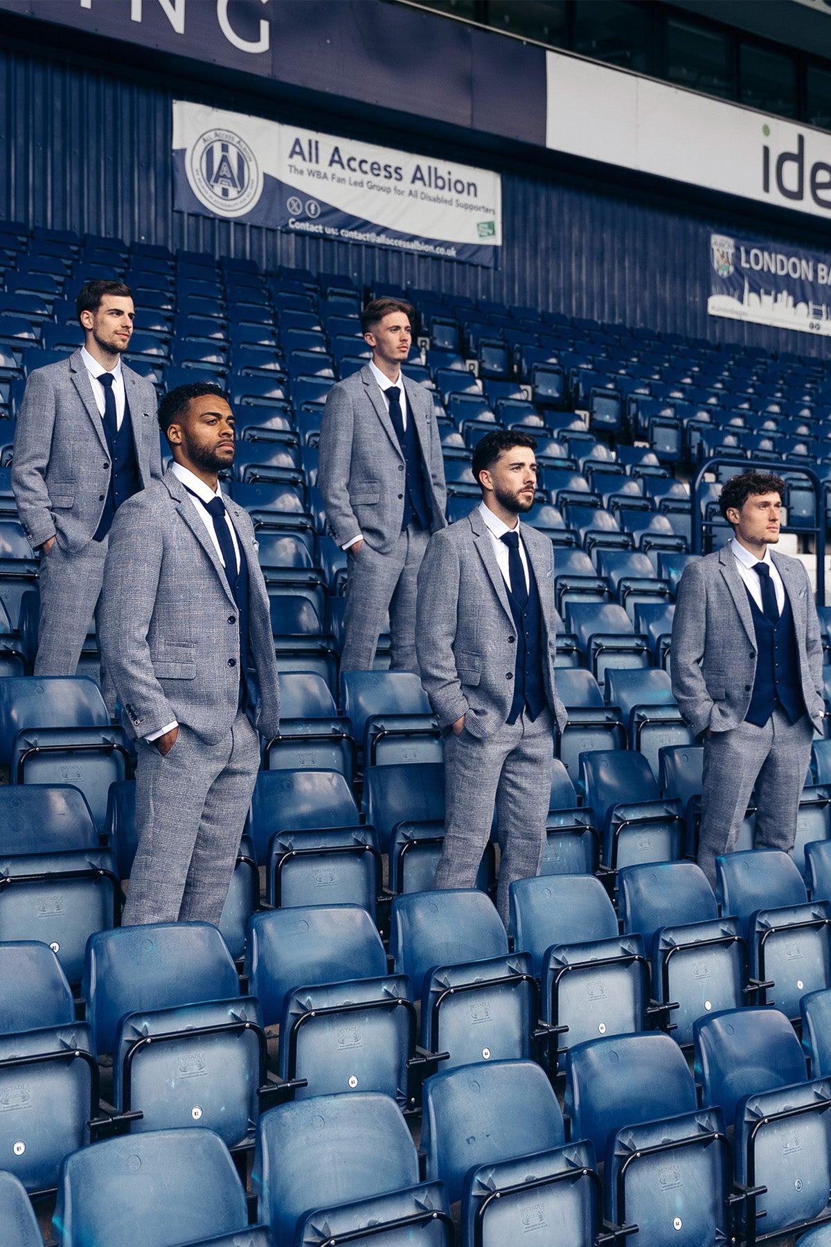 Five men in matching marcdarcy Official West Bromwich Albion F.C. Club Suits (KRIS Tweed Suit with Kelvin Navy Waistcoat) stand among empty blue stadium seats, a sign and dark blue wall behind them, looking ahead with serious expressions.