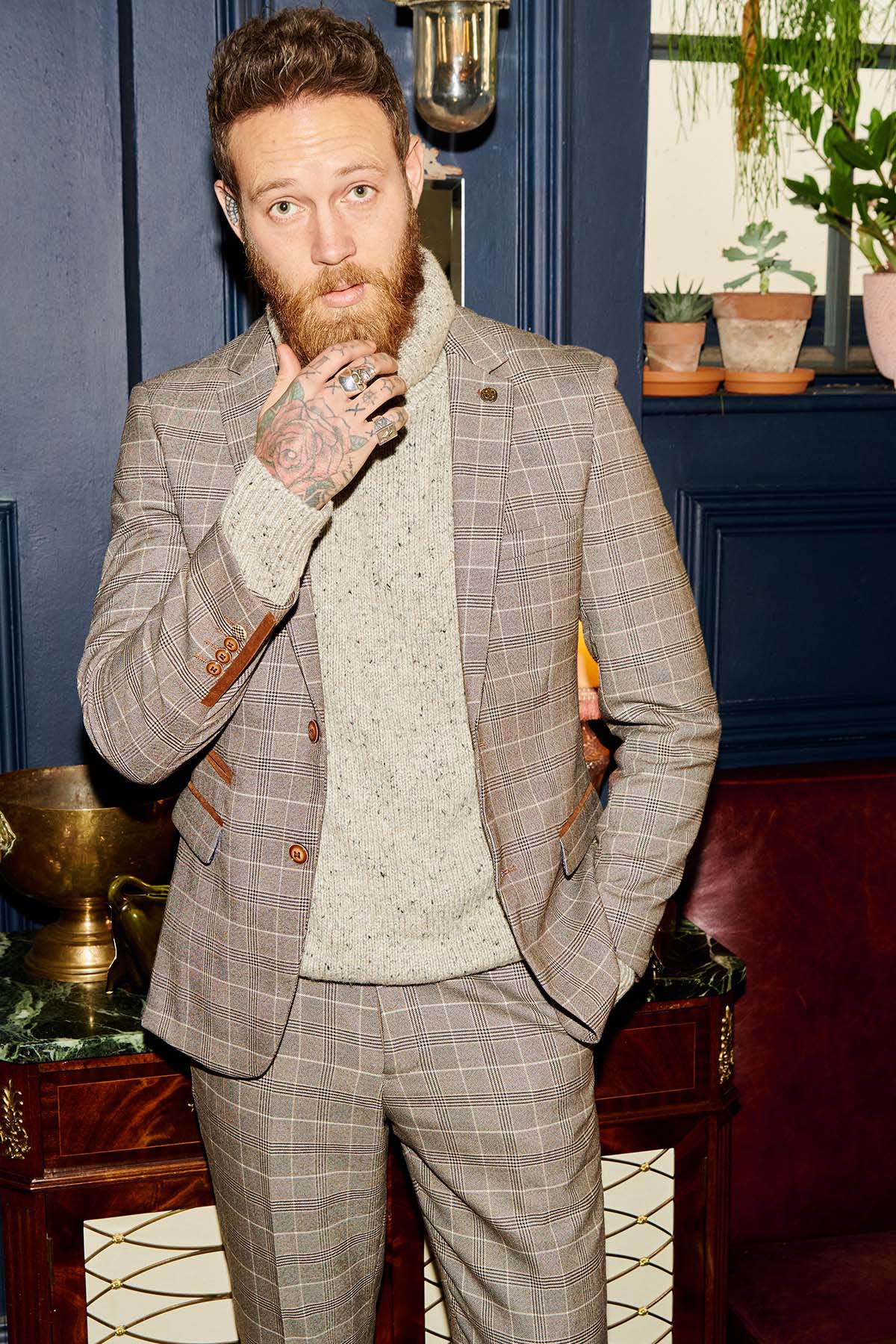 A bearded, tattooed man wears the marcdarcy RAY - Tan Check Two Piece Suit over a light knit sweater, posing indoors with his hand on his chin. Potted plants and a dark blue wall set a stylish officewear scene.