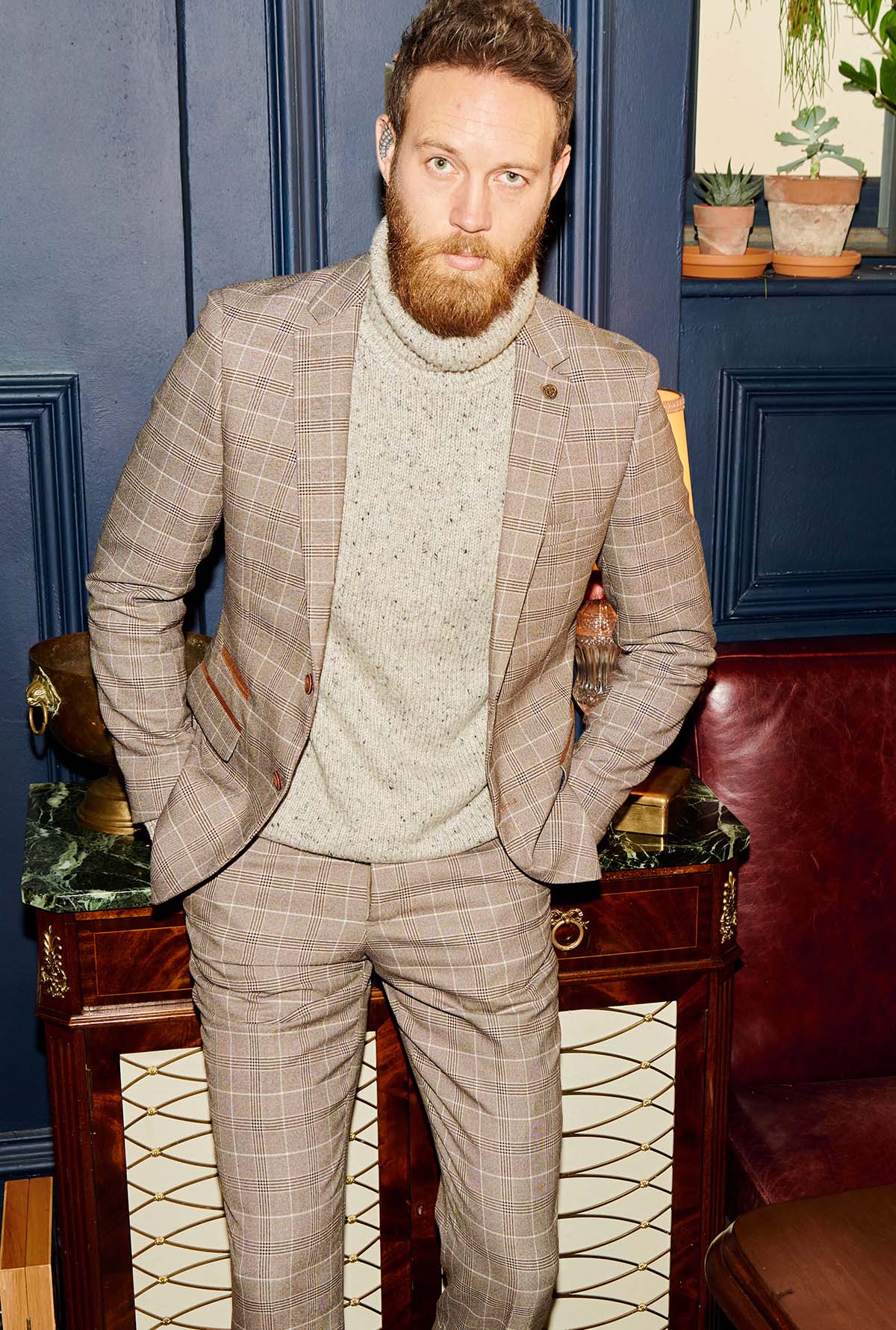 A man with a beard wears the marcdarcy RAY - Tan Check Two Piece Suit and a light gray turtleneck, standing indoors by a dark wood cabinet with potted plants against dark blue paneled walls.