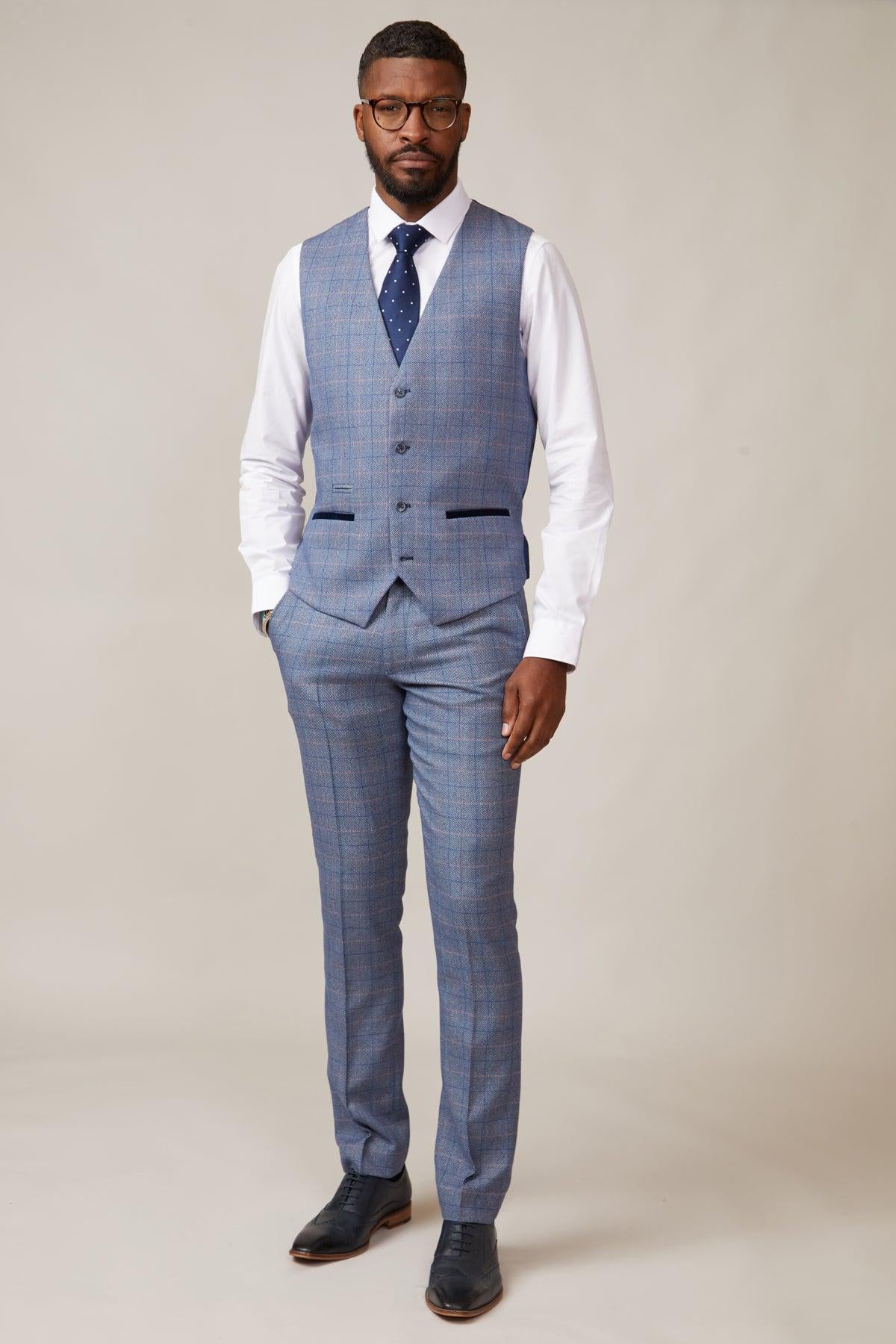 A man stands against a plain background wearing the marcdarcy HILTON Blue Tweed Check Single Breasted Waistcoat, a white dress shirt, navy polka dot tie, black dress shoes, and glasses. He has short hair and a beard.