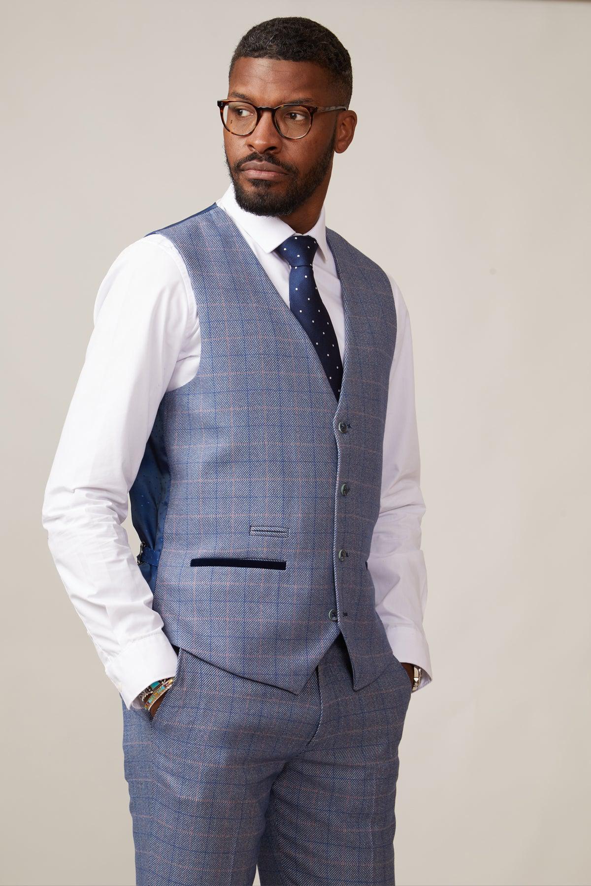 A Black man wearing the marcdarcy HILTON Blue Tweed Check Single Breasted Waistcoat, a white shirt, navy polka dot tie, and glasses, with his hands in his pockets.